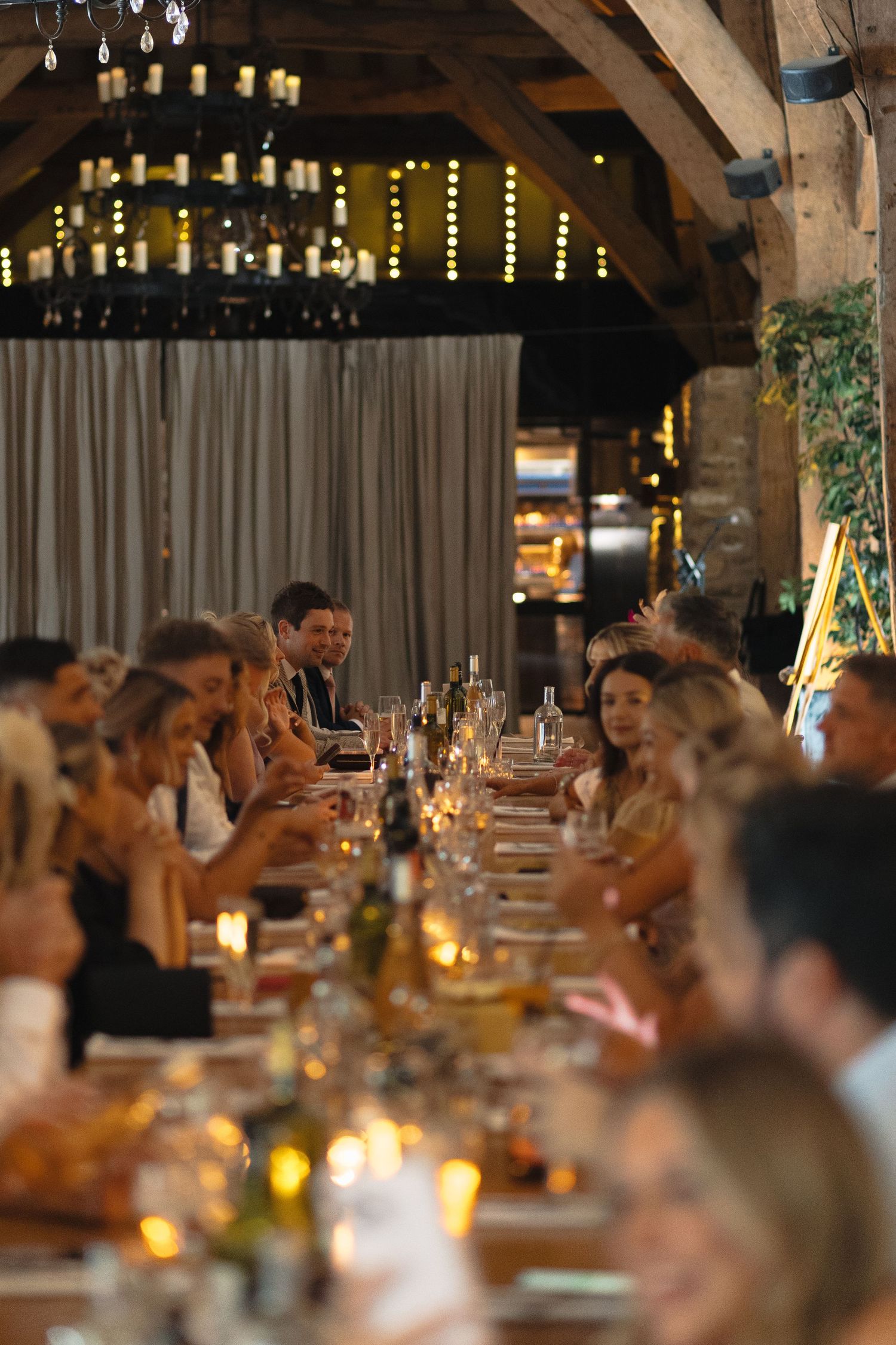 Long banquet table filled with guests enjoying a wedding reception dinner under wooden beams.