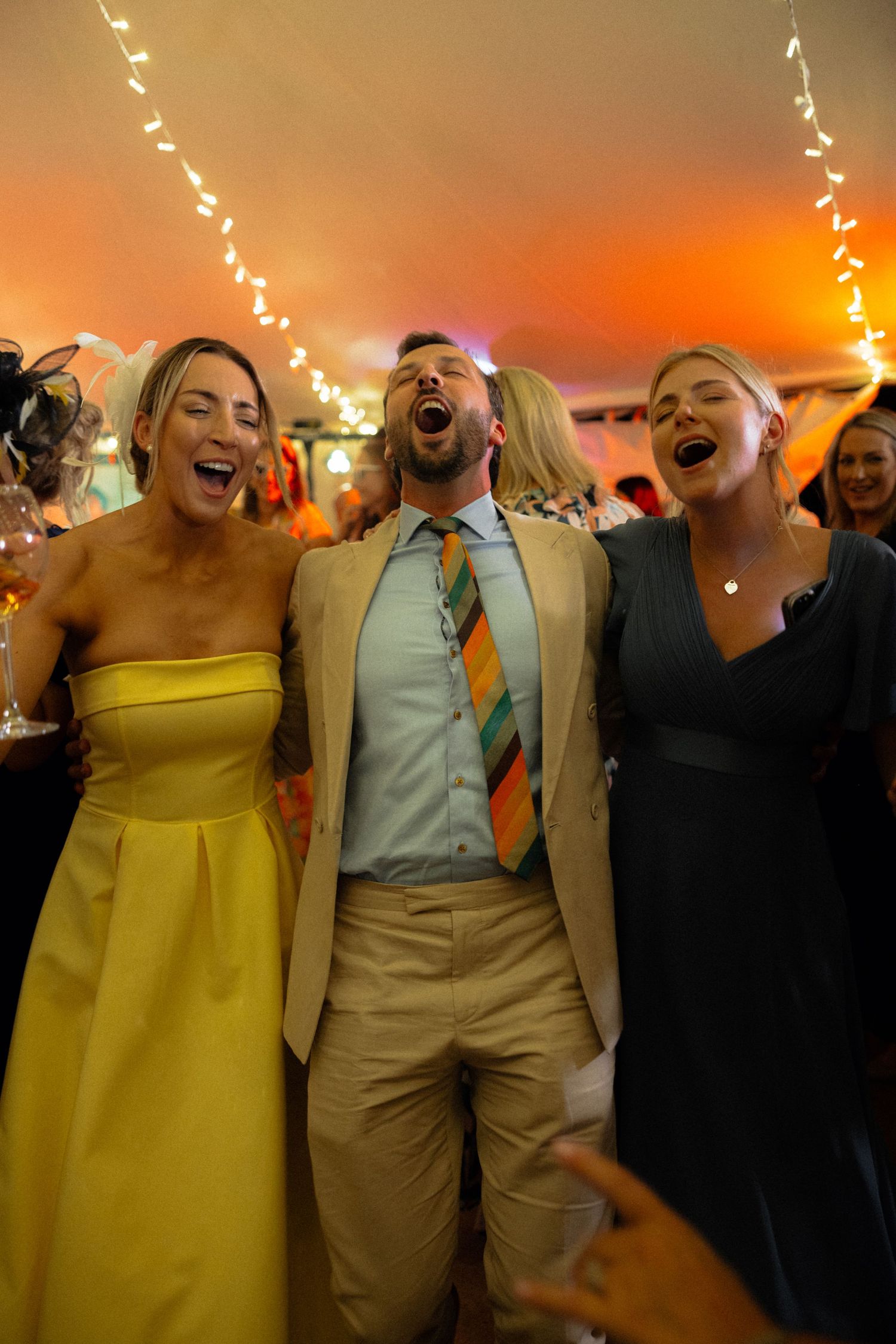 Wedding party guests singing and laughing together under string lights at reception.