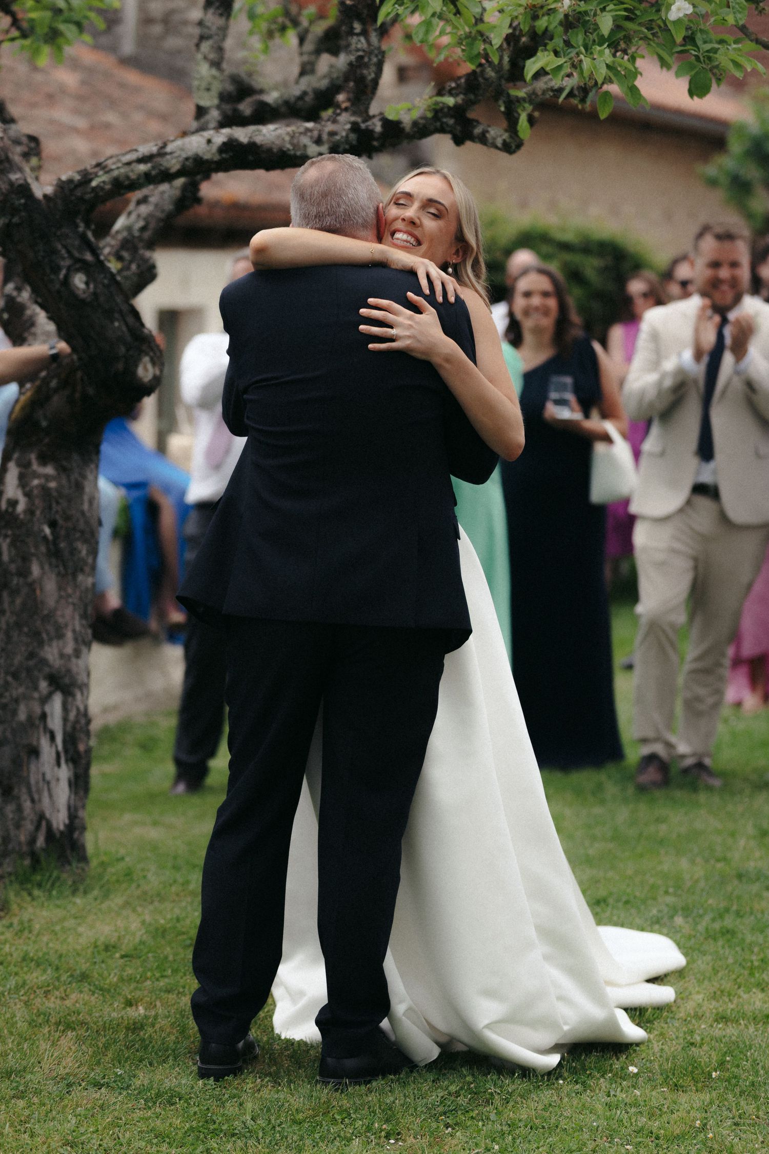 A bride in a white dress embraces someone during an outdoor wedding celebration.
