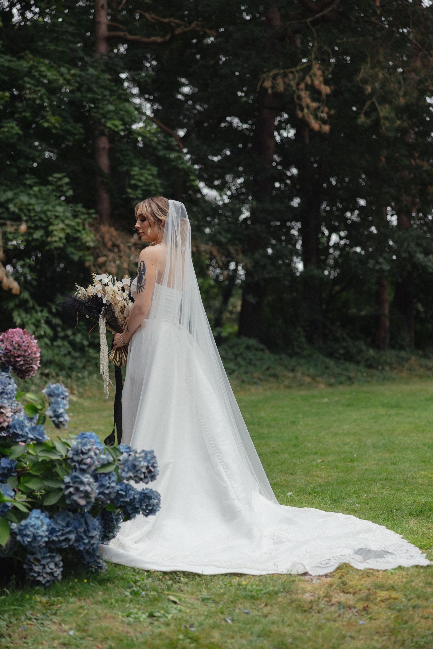 Wedding dress train trailing elegantly across grass with blue hydrangeas in the foreground.