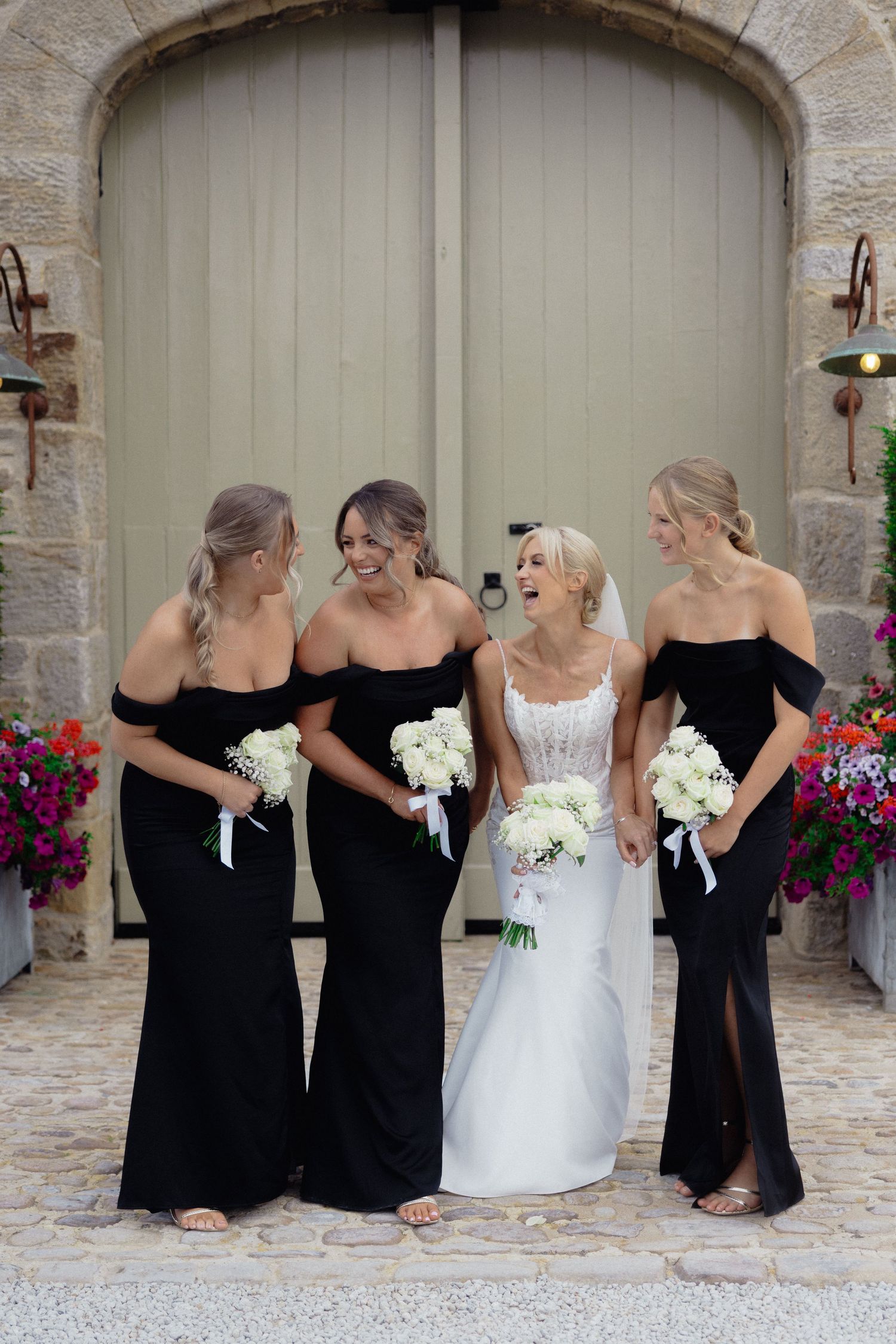 Bridesmaids in black dresses and bride in white pose together against stone archway.