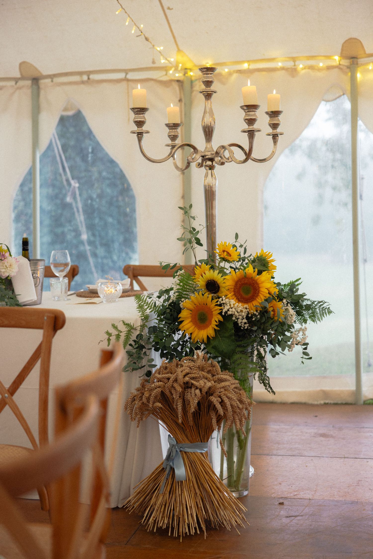 Rustic wedding tent decor featuring sunflower arrangements and wheat bundles under string lights.