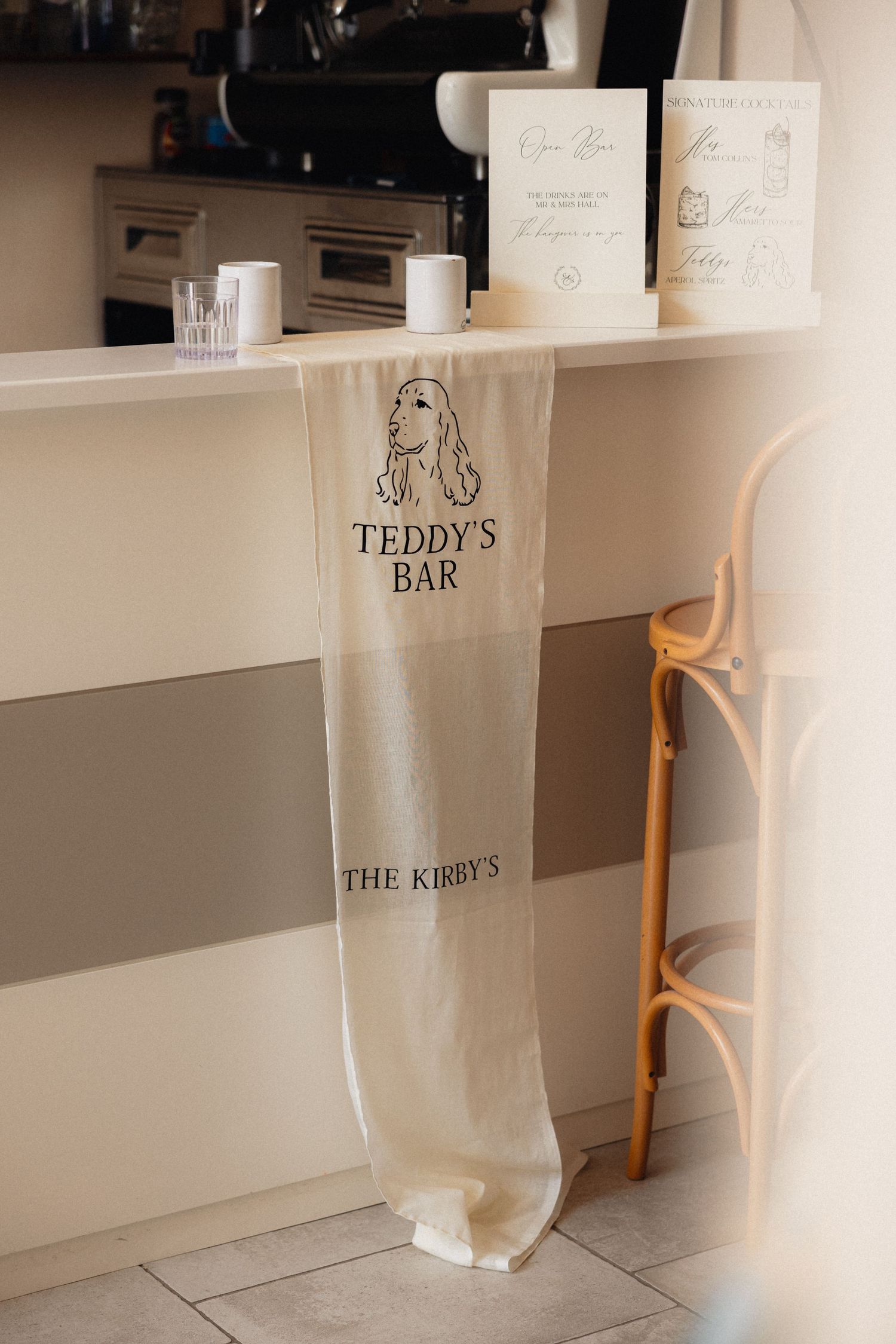 Wedding venue setup with white decor and signs including 'Teddy's Bar' displayed on shelving.