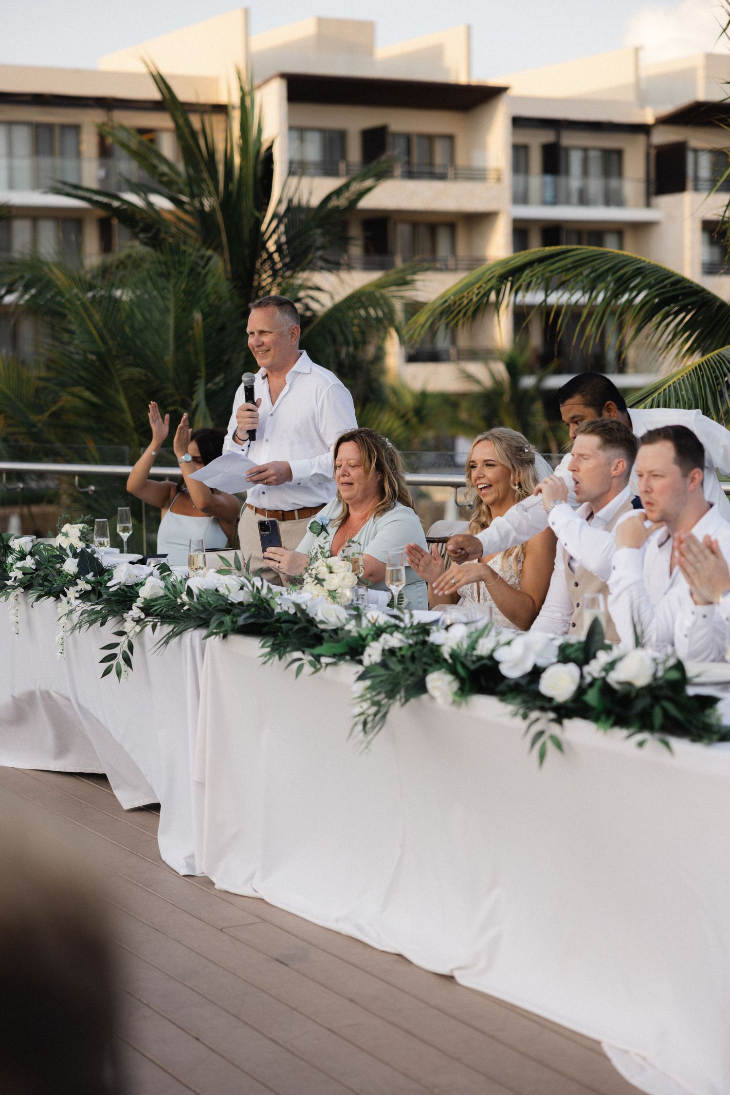 Wedding party seated at white-decorated head table with tropical palm backdrop.