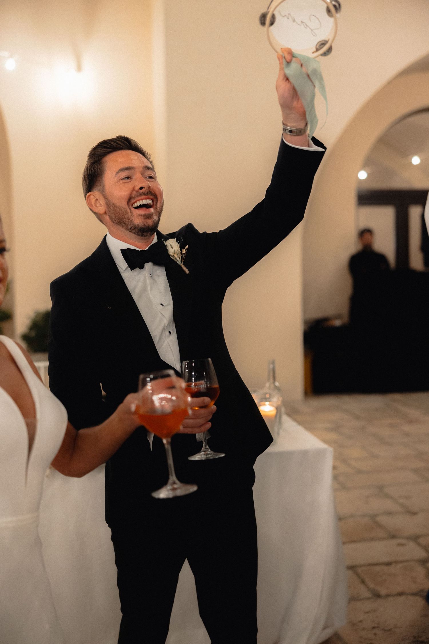 Wedding guests in formal attire celebrate joyfully with raised arms and drinks at an elegant indoor reception.