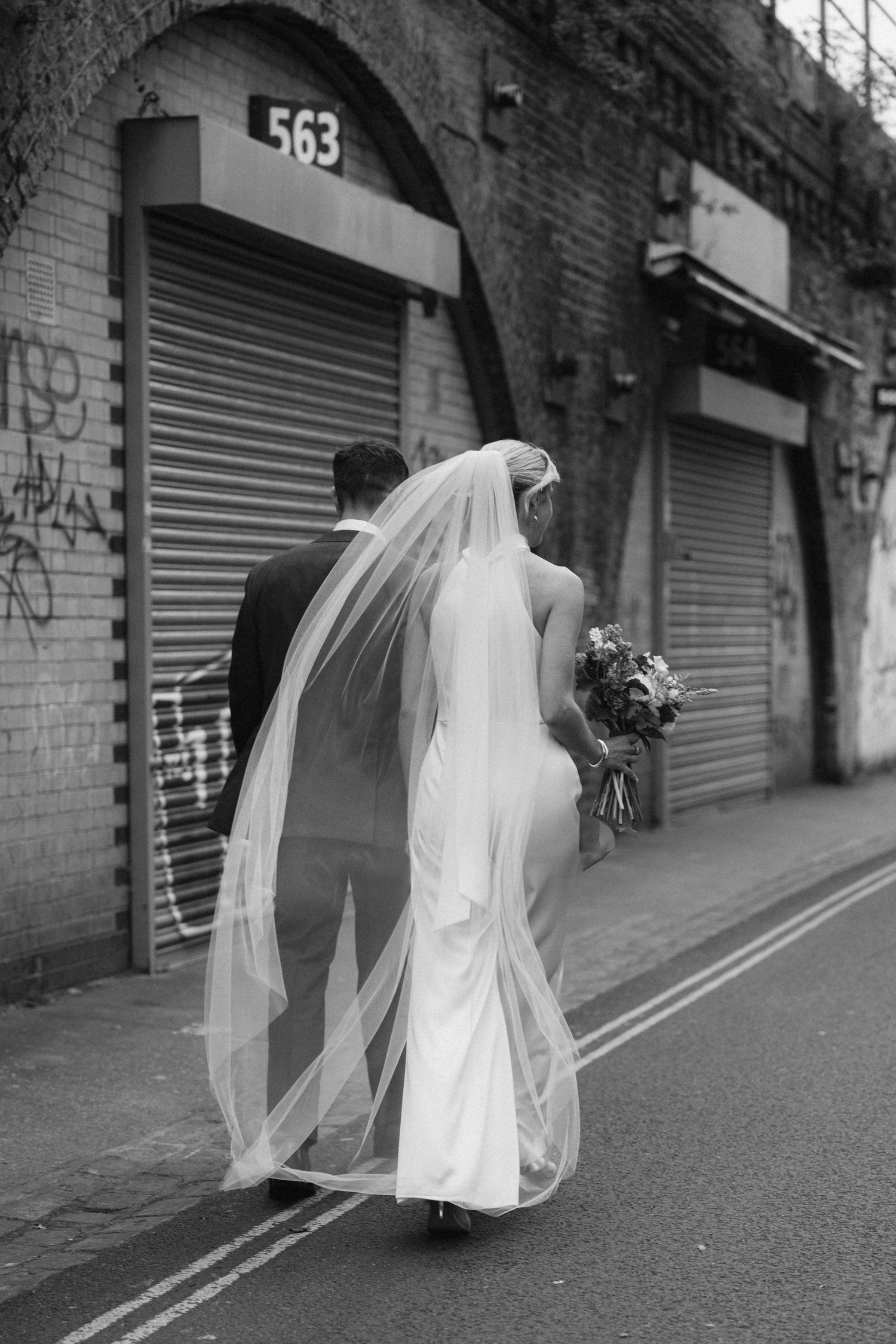 Black and white photo of bride with flowing veil walking along urban street with shuttered storefronts.