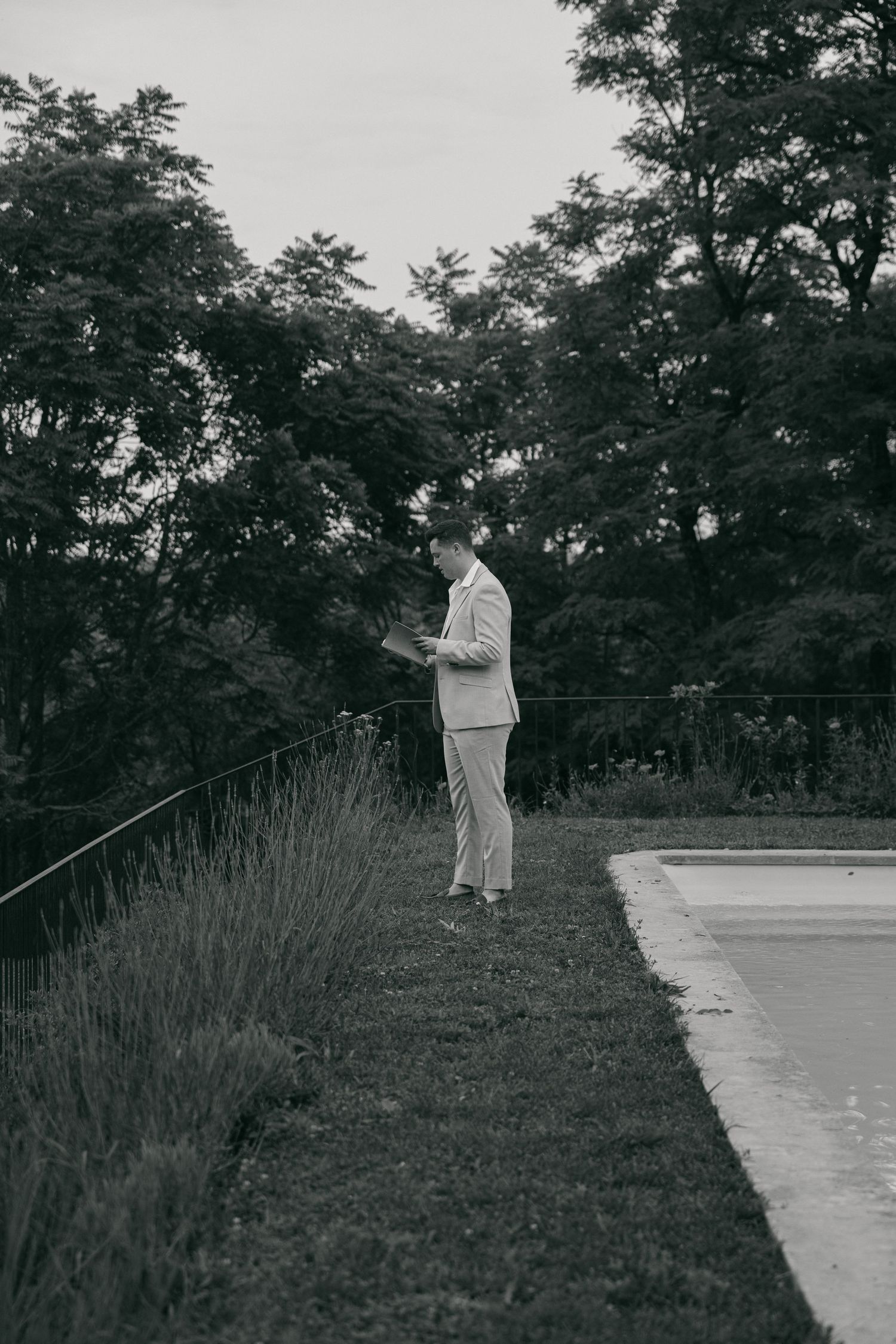 A figure in light colored clothing stands on a concrete path near tall grass in a black and white photograph.