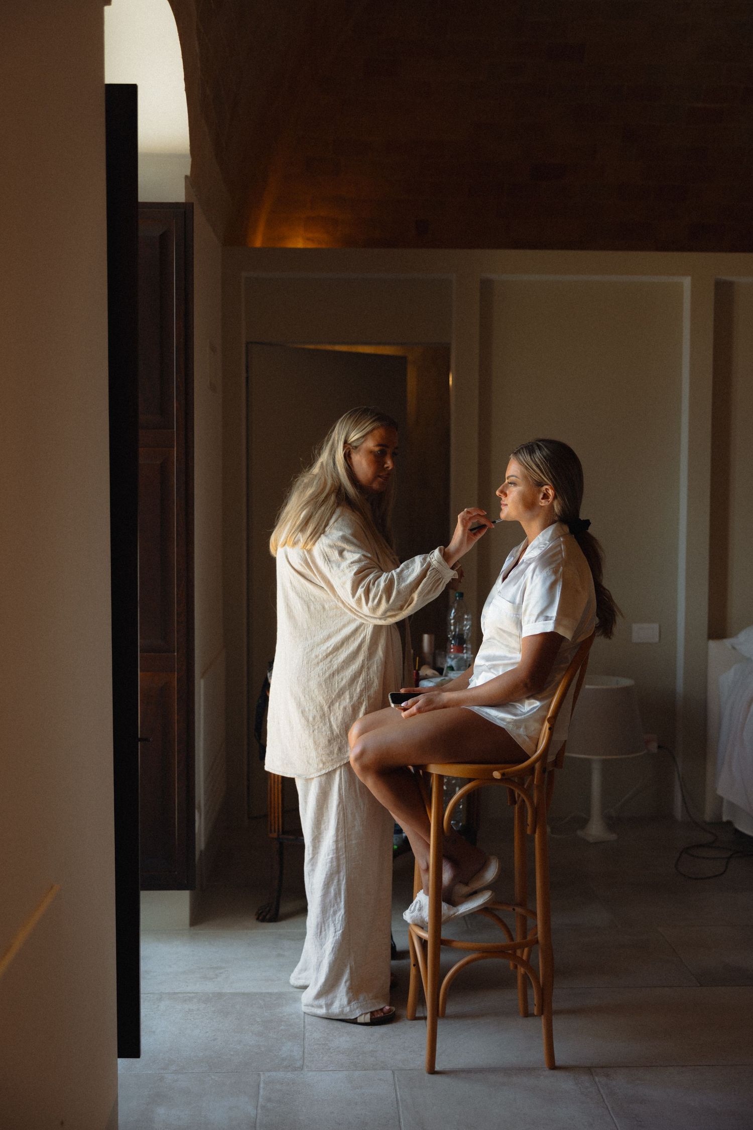 Makeup artist applying cosmetics to a person seated in a wooden chair.