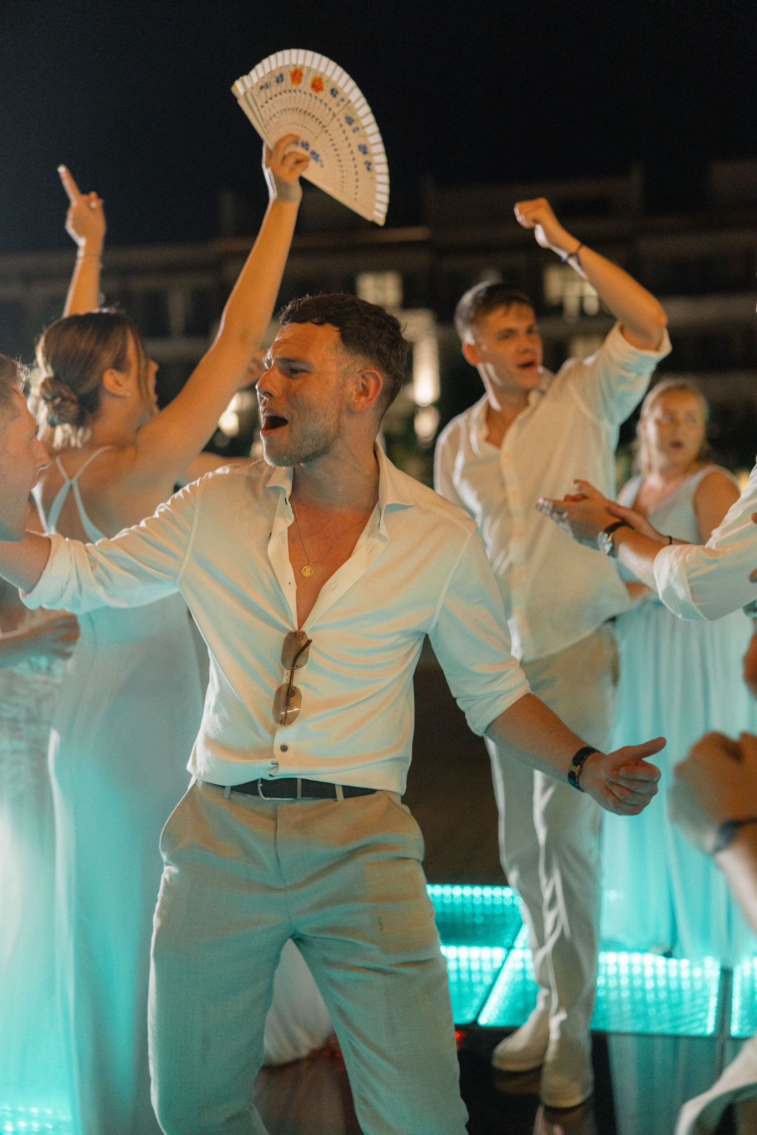 Energetic dancers in white outfits perform at a wedding reception with dramatic lighting.