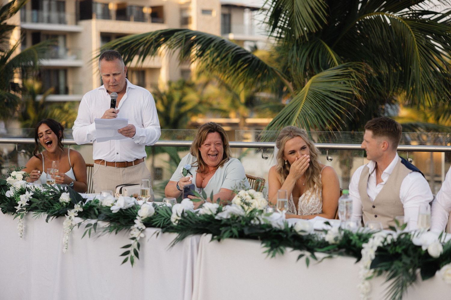 Wedding reception head table decorated with greenery and white linens at tropical resort.