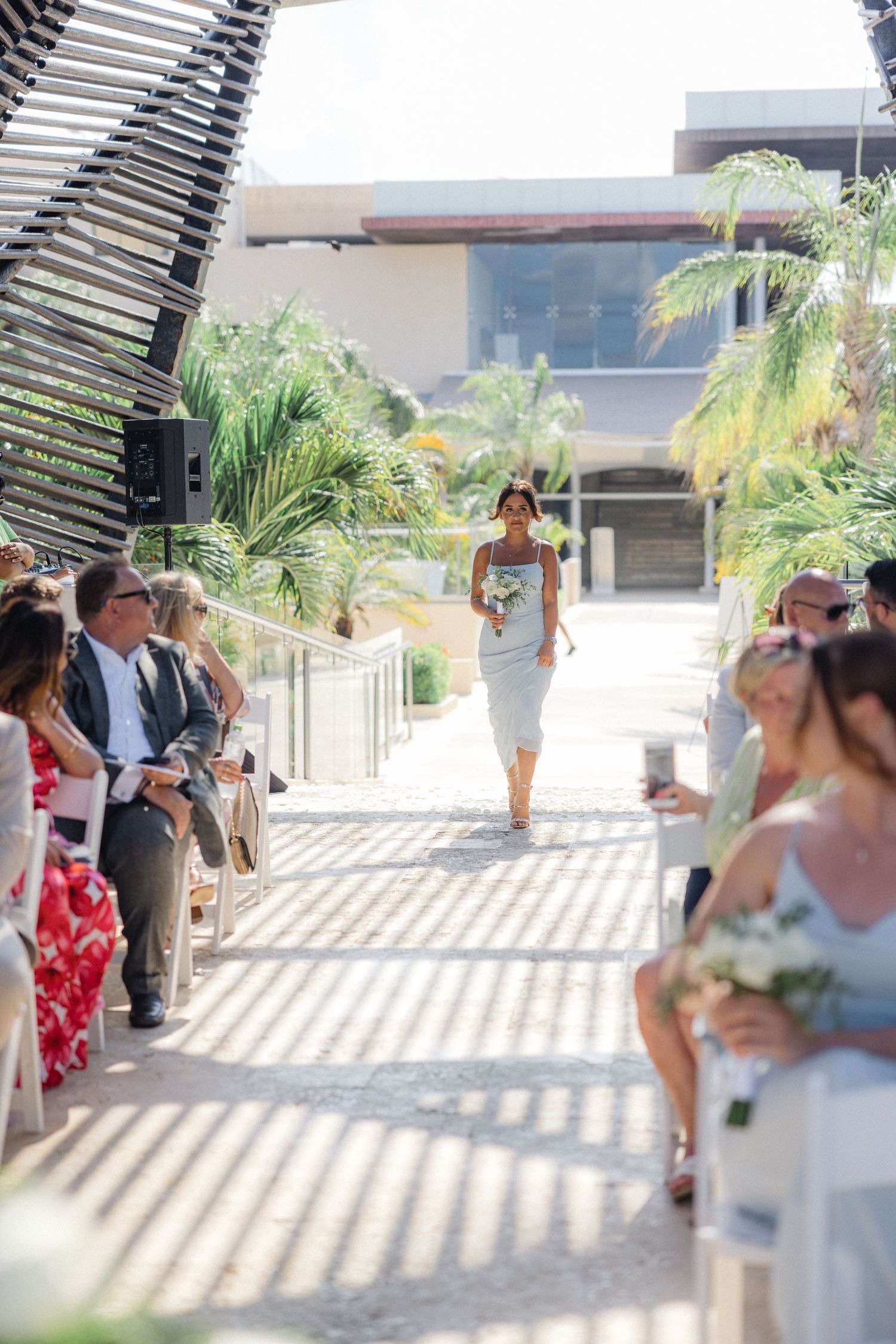 Outdoor wedding venue with palm trees and guests seated under a slatted roof structure.