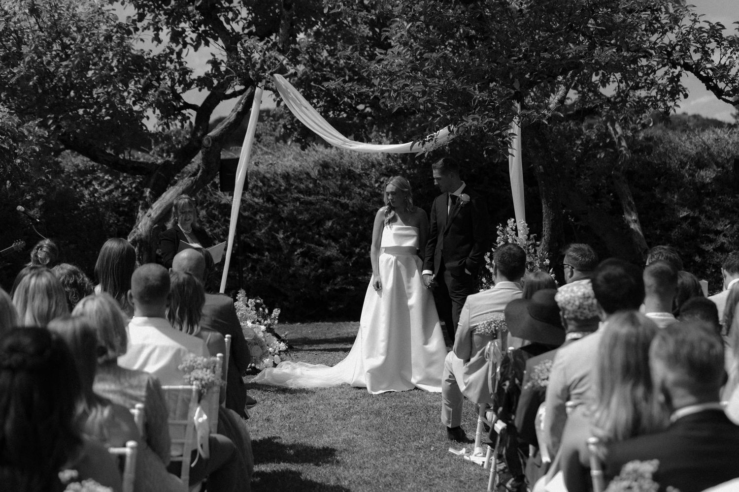 A black and white photo of an outdoor wedding ceremony with guests seated under draped fabric decor.