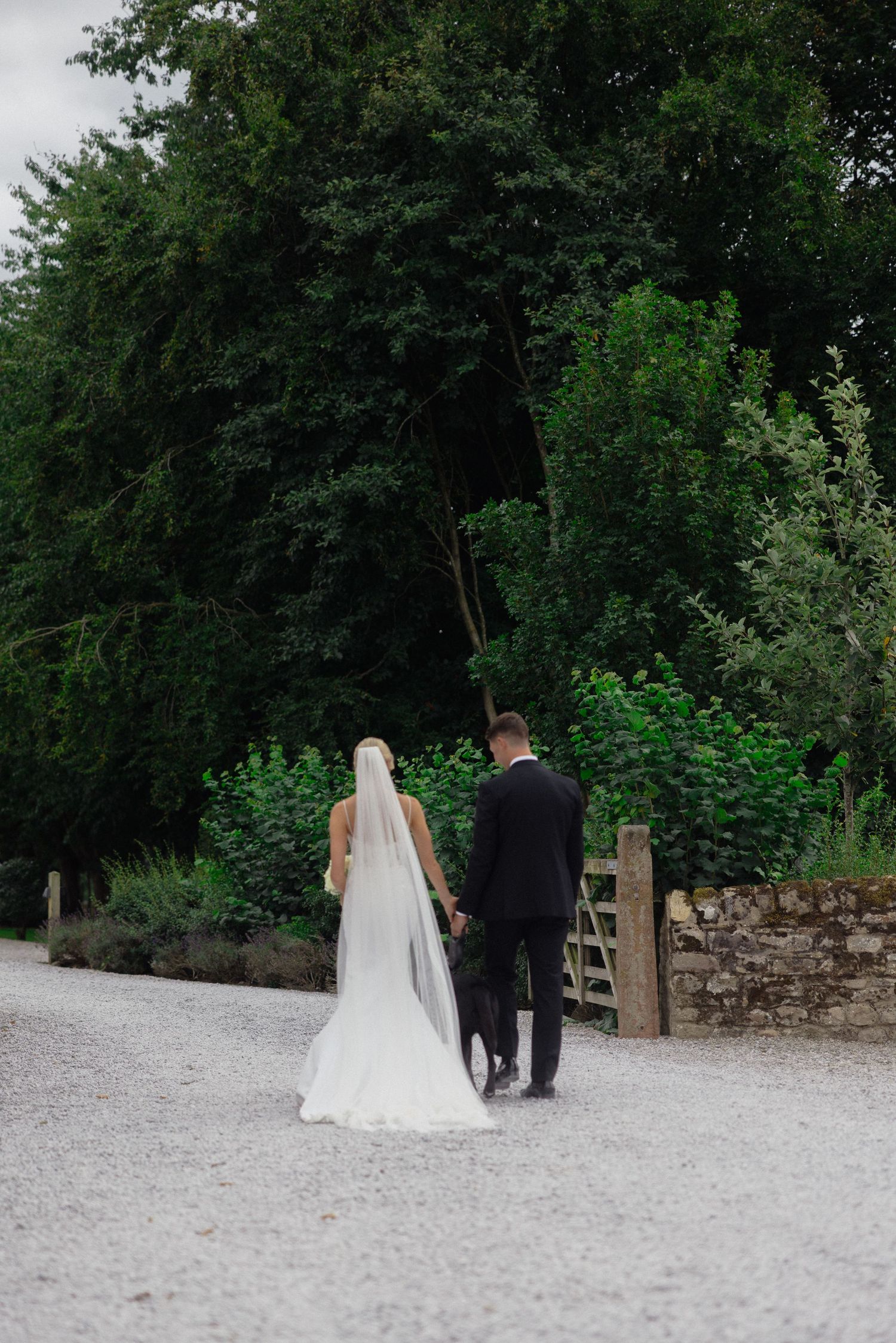 Bride and groom walking away on gravel path surrounded by green trees.