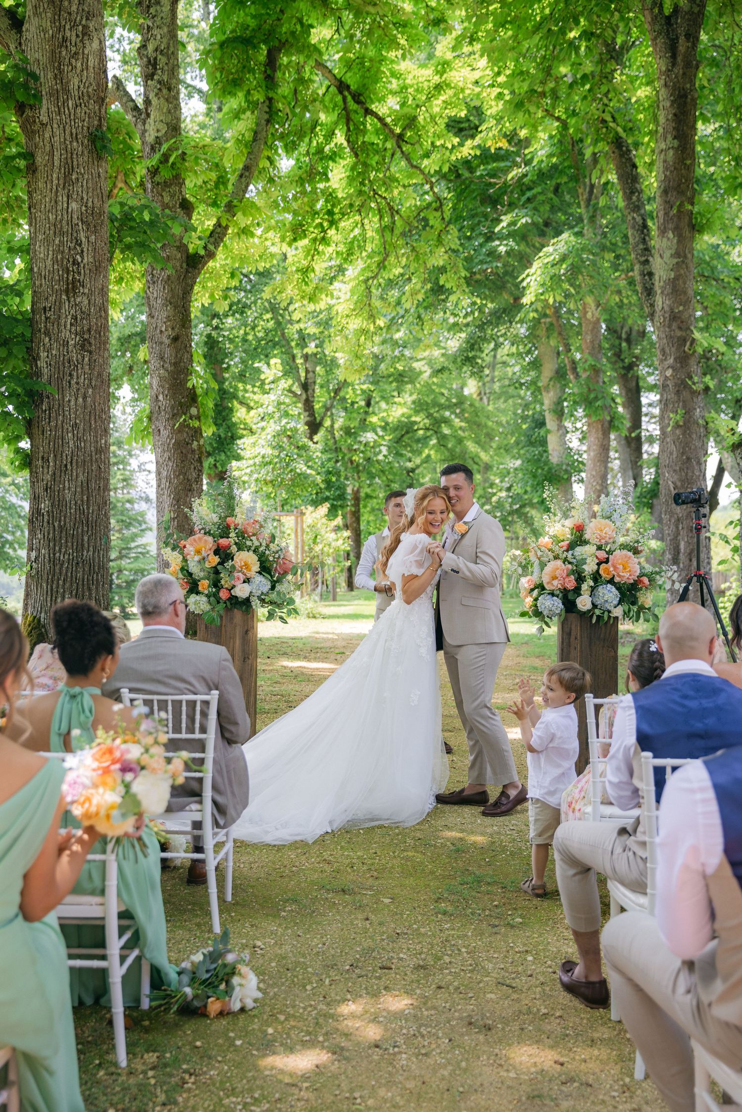 Wedding guests seated in white chairs beneath a canopy of trees during an outdoor ceremony.