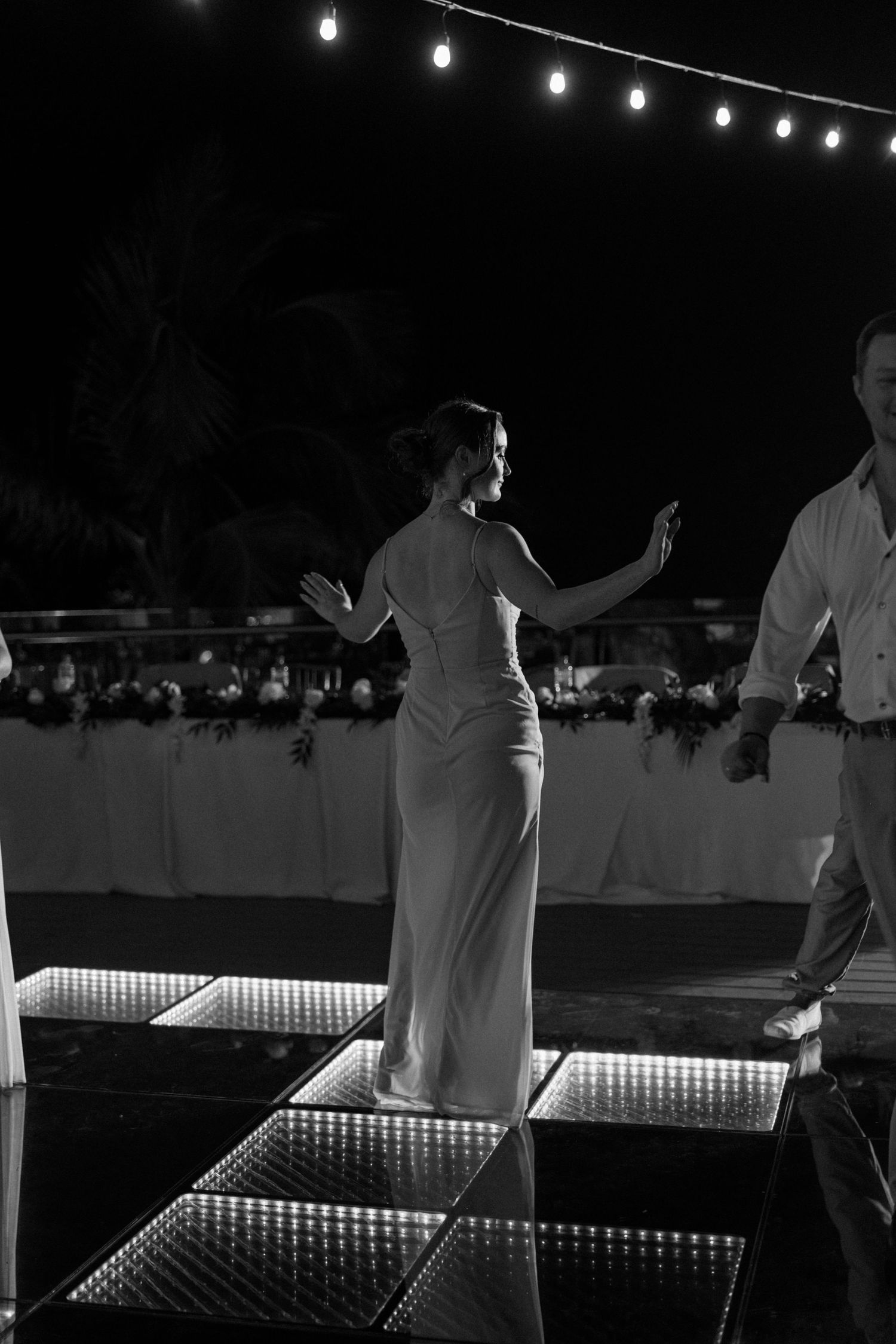 Couple dancing on an illuminated dance floor at night with string lights overhead.
