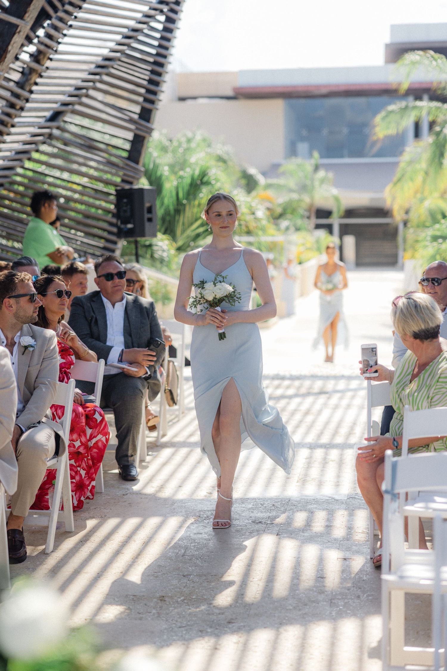 Wedding guests seated in white chairs under a slatted pergola casting dramatic shadows.
