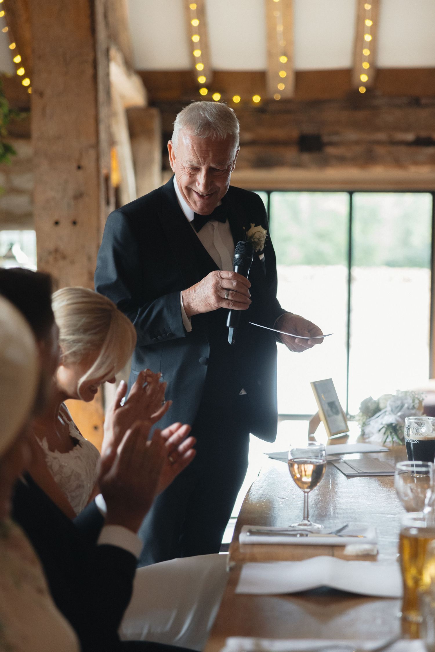 Wedding reception guests enjoying speeches in a rustic venue with natural lighting.