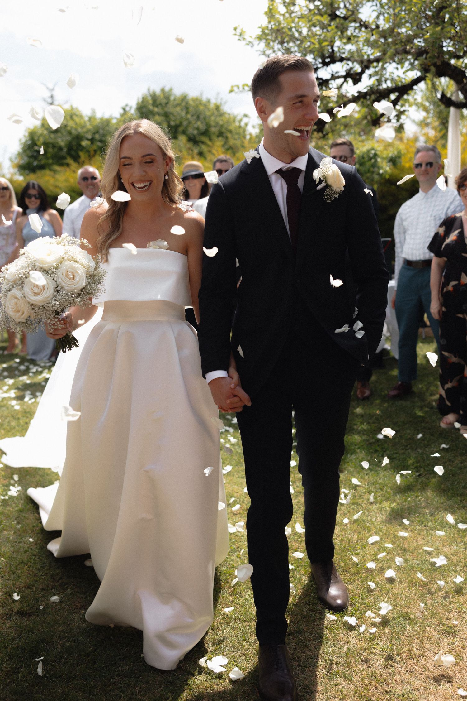 Newlyweds walk through shower of flower petals during outdoor garden ceremony.
