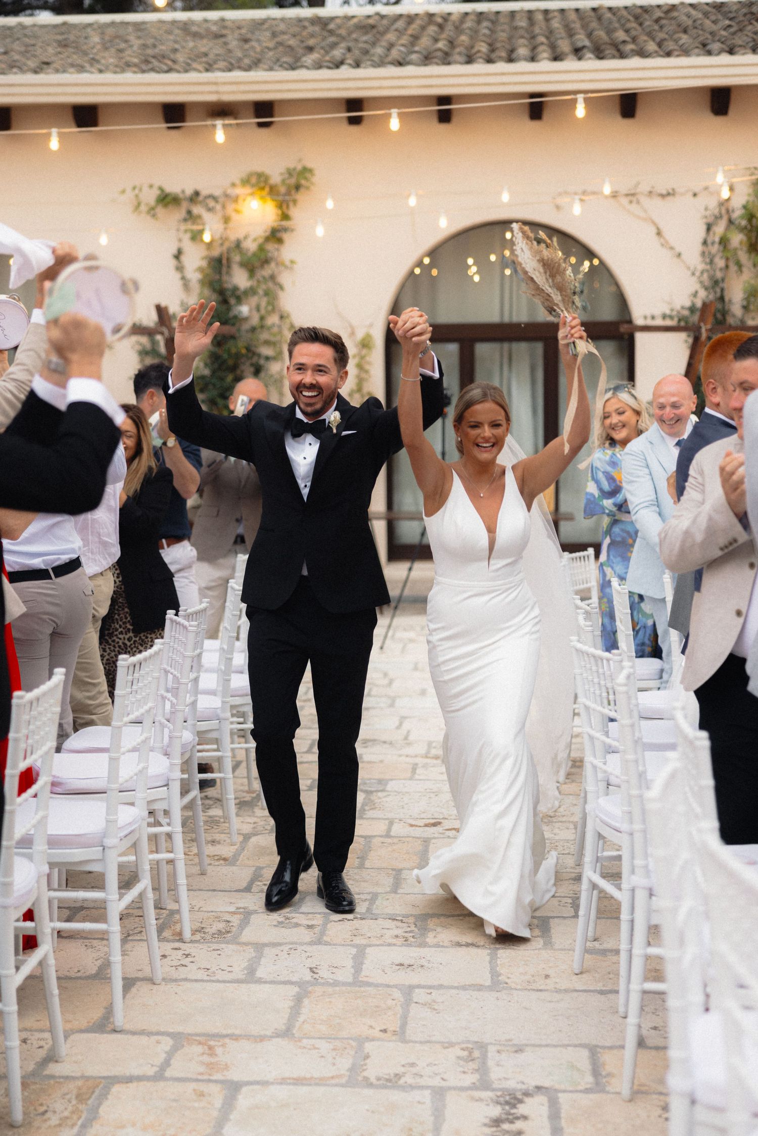 Wedding party celebrates with raised arms between rows of white chairs at an outdoor venue.