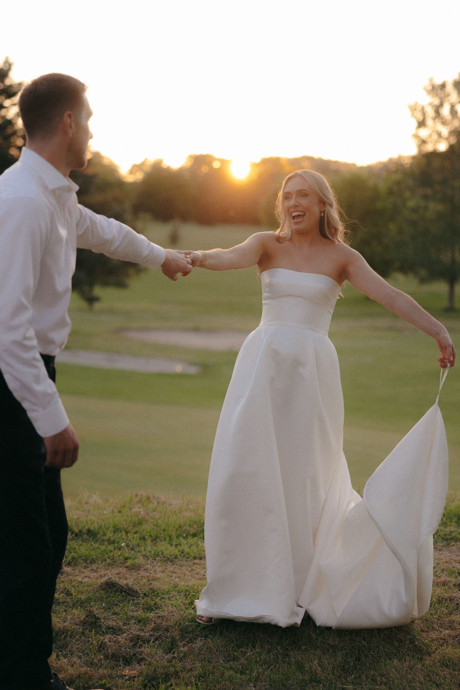 Wedding couple dances and twirls in a sunset-lit field wearing formal attire.