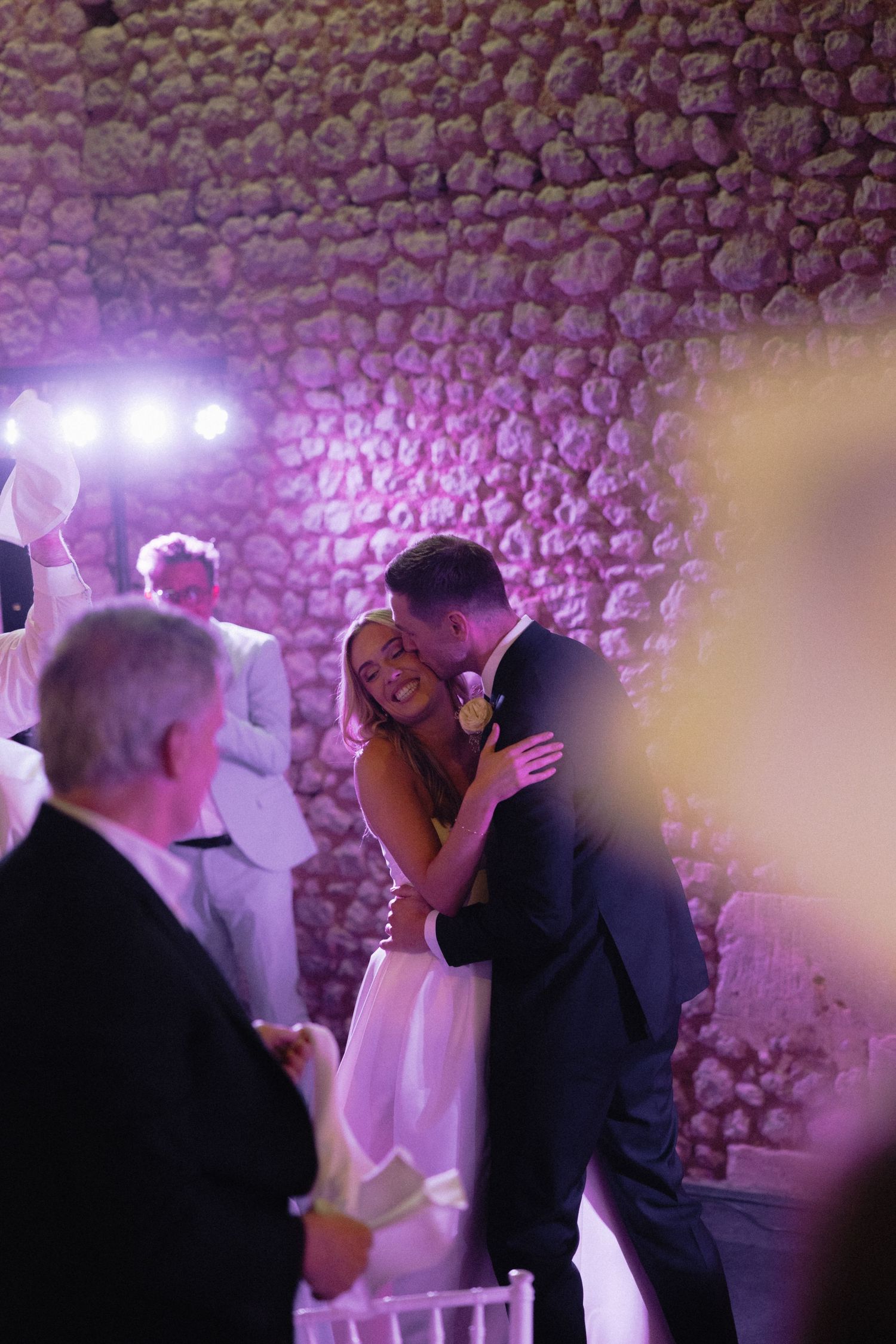 Couples dancing at a wedding reception with purple lighting and stone wall backdrop.