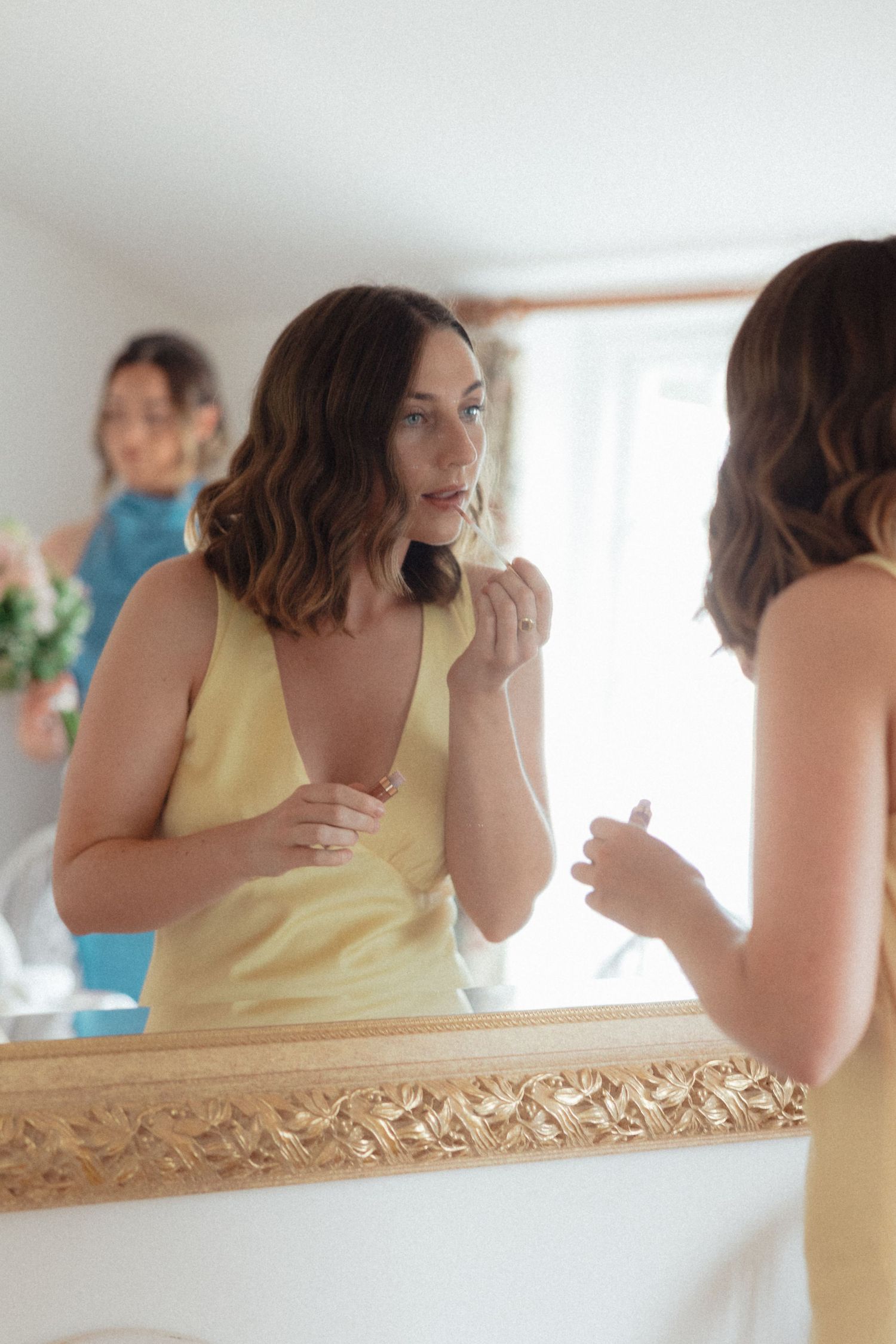 Yellow-dressed attendants reflected in ornate gold mirror during wedding preparations.