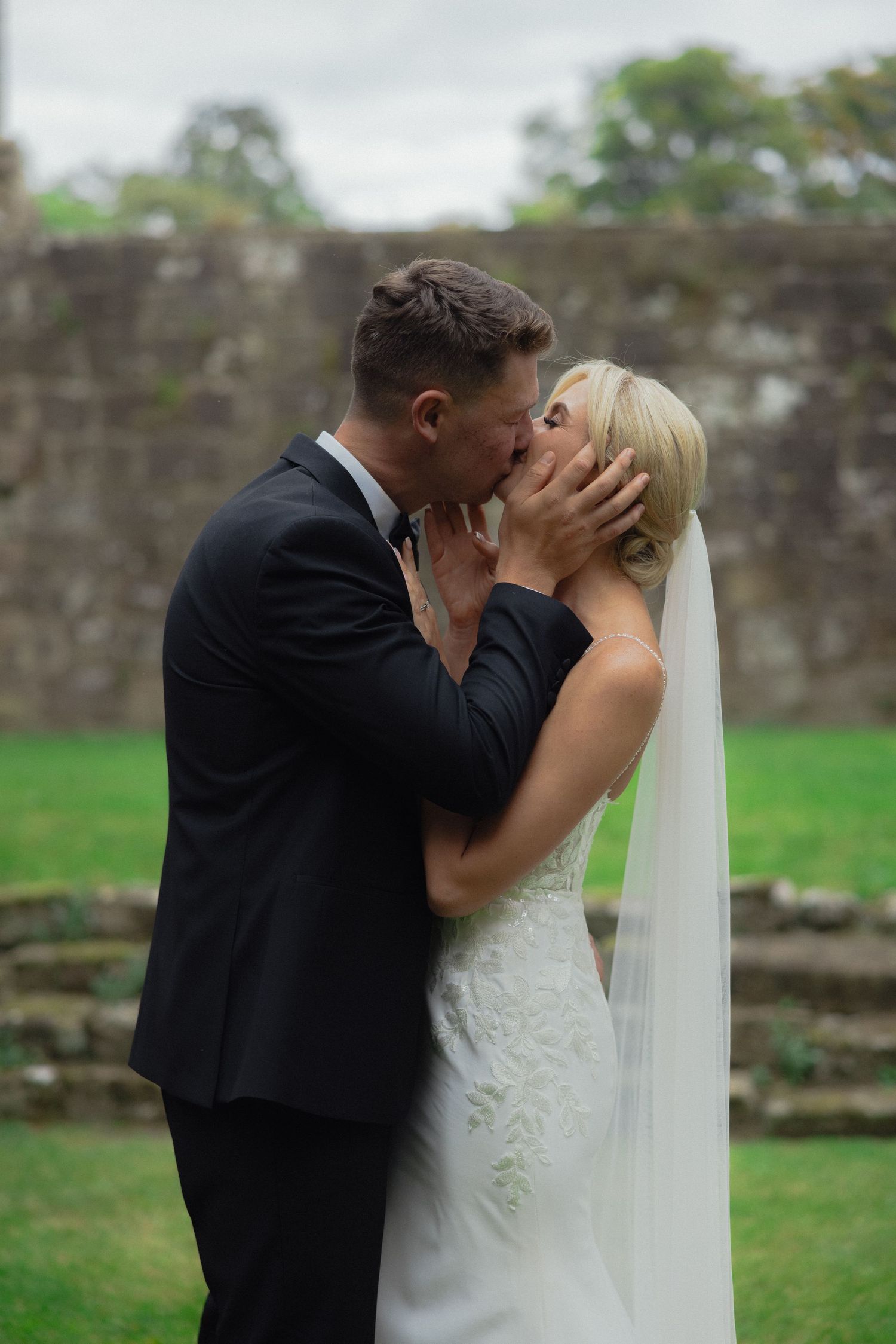 Romantic wedding kiss captured against historic stone wall backdrop.