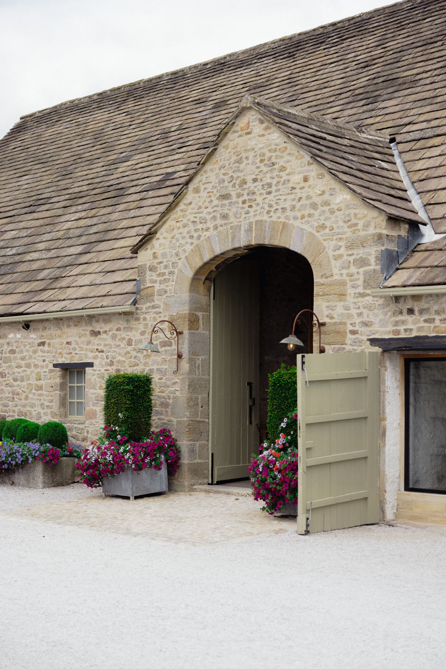 Stone barn entrance with green door and colorful flower boxes under a slate roof.