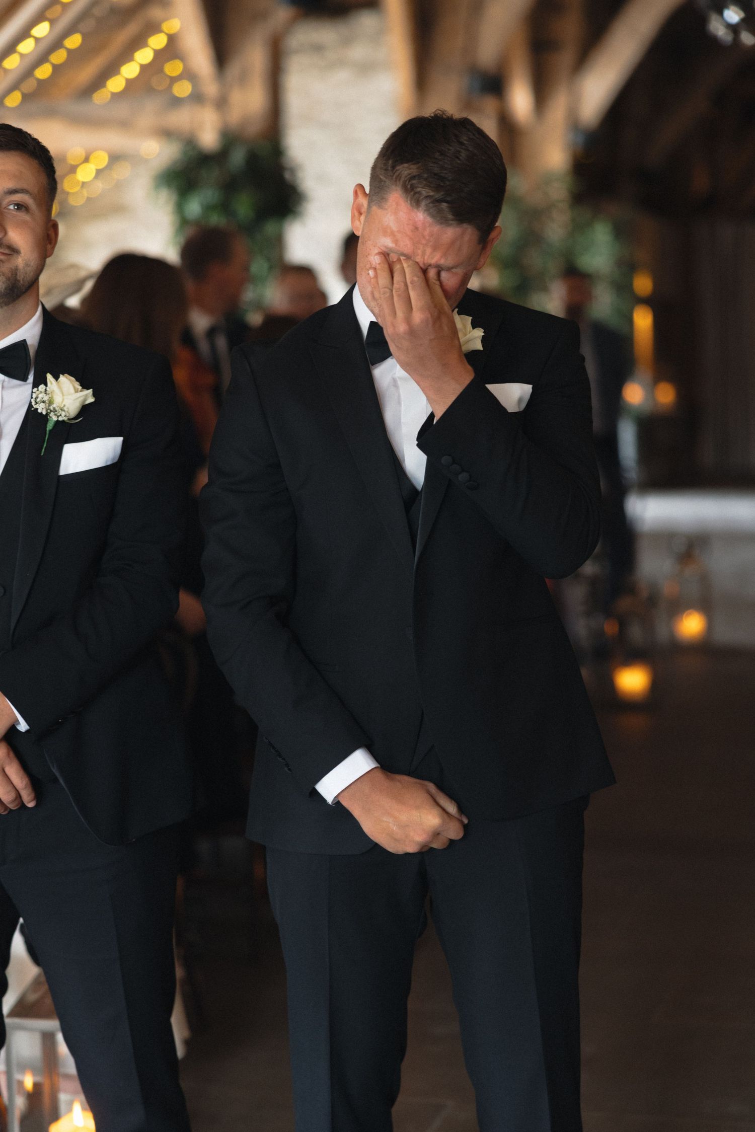Emotional groomsmen react during wedding ceremony in rustic barn setting.