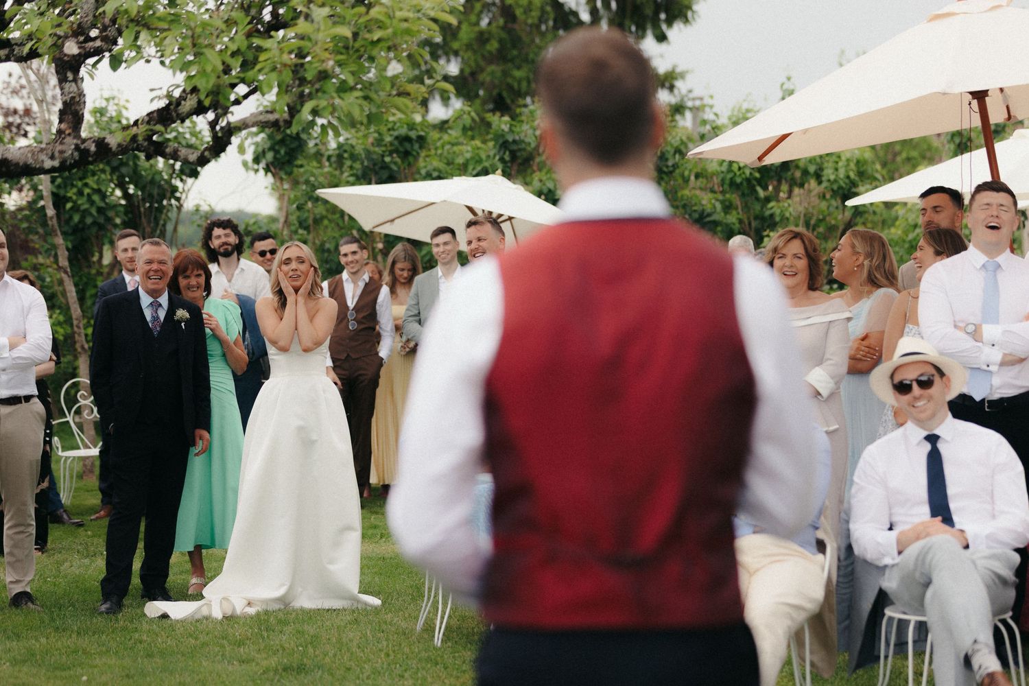 Outdoor wedding reception with guests gathered under trees and white umbrellas.