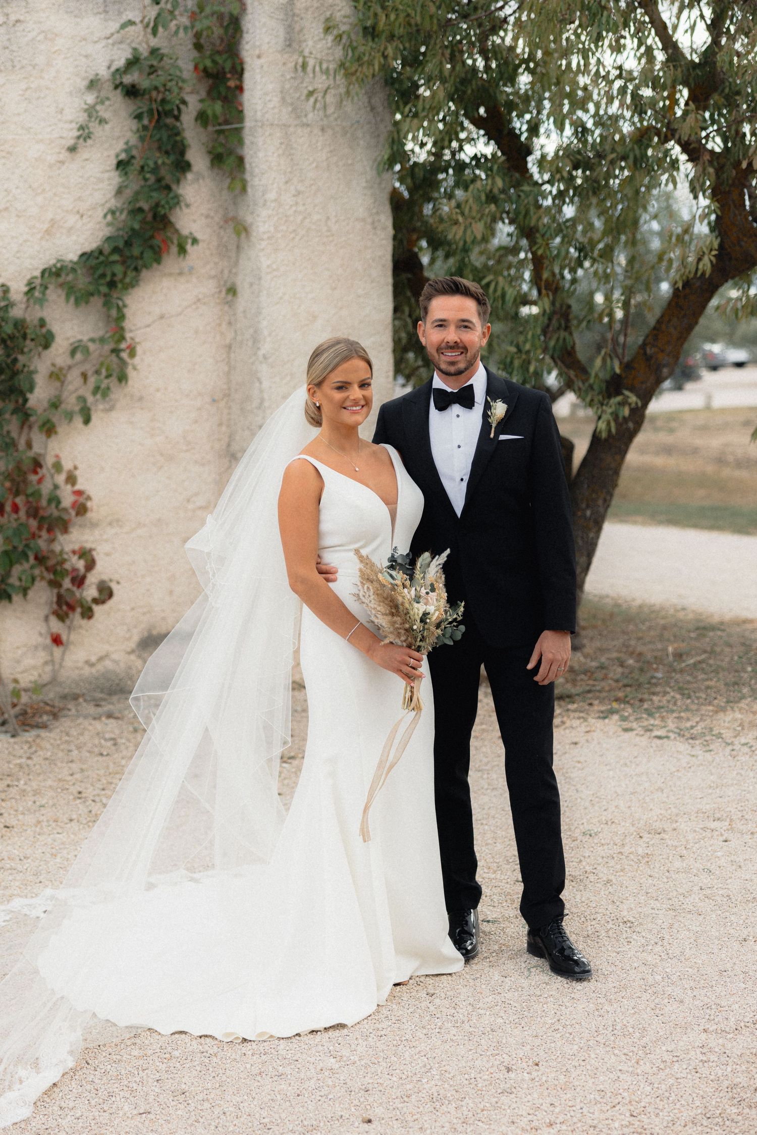 A couple poses for wedding photos against a rustic wall with climbing vines and greenery.