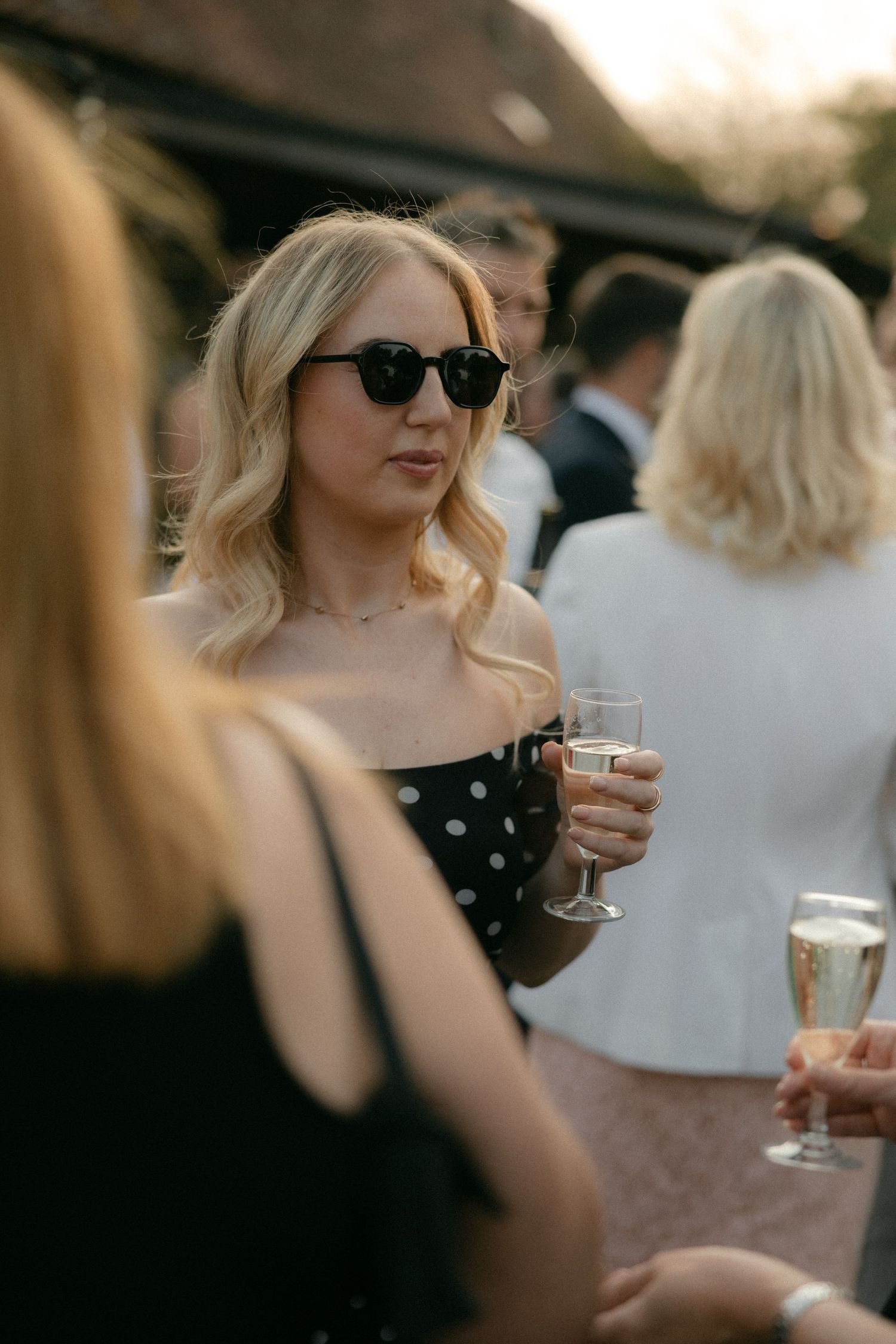 A woman in sunglasses and polka dot dress enjoys drinks with friends at an outdoor gathering.