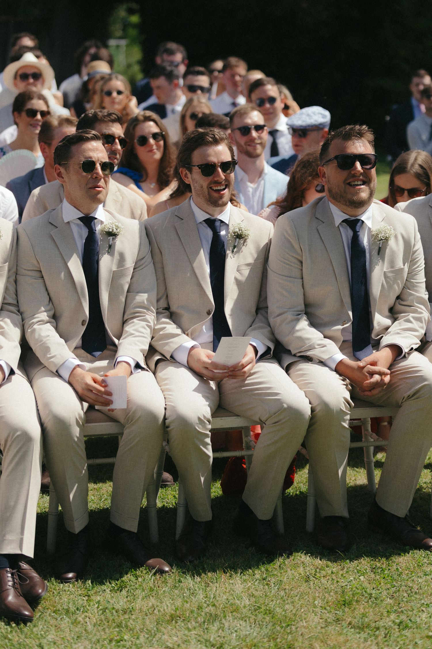 Groomsmen in light beige suits sitting on outdoor bench during wedding ceremony.