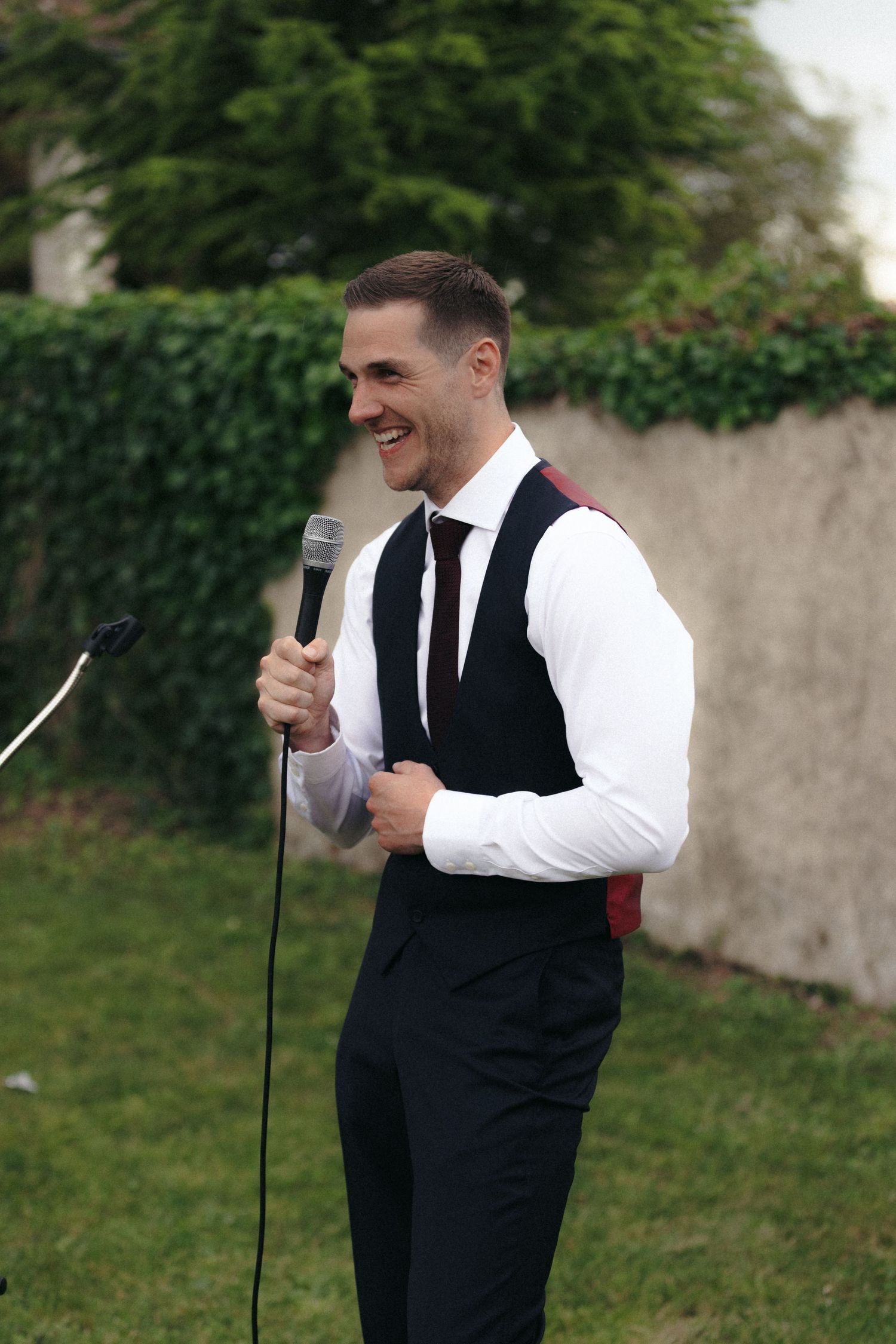 Speaker in formal vest and tie holds microphone during outdoor event.
