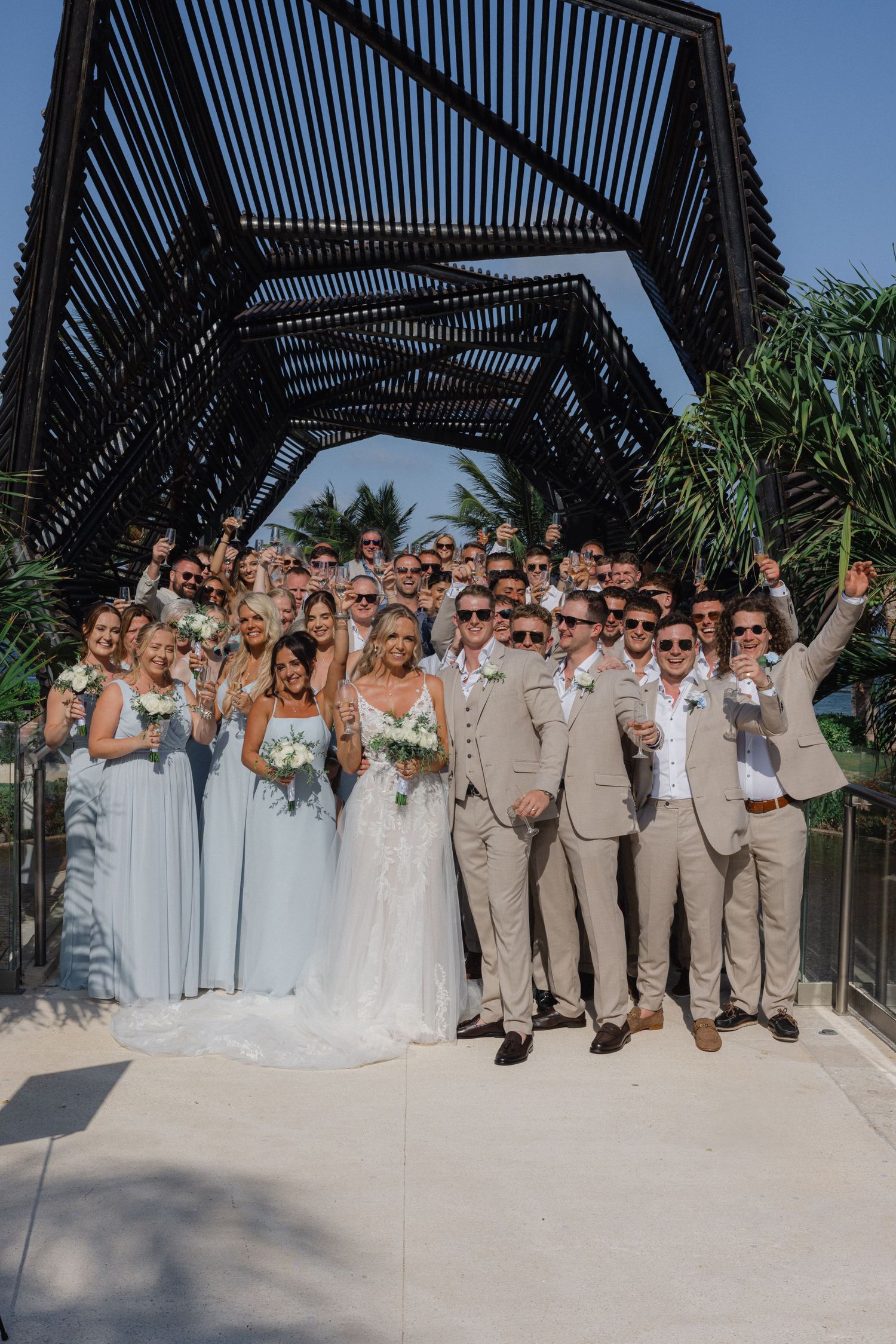 Wedding party poses for group photo under modern black pergola at tropical venue.