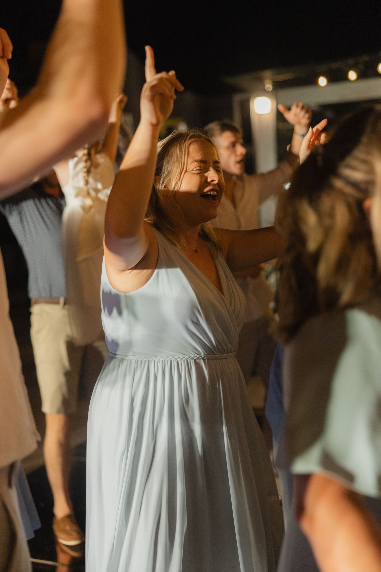 Wedding guest in light blue dress dances with hands raised during reception celebration.
