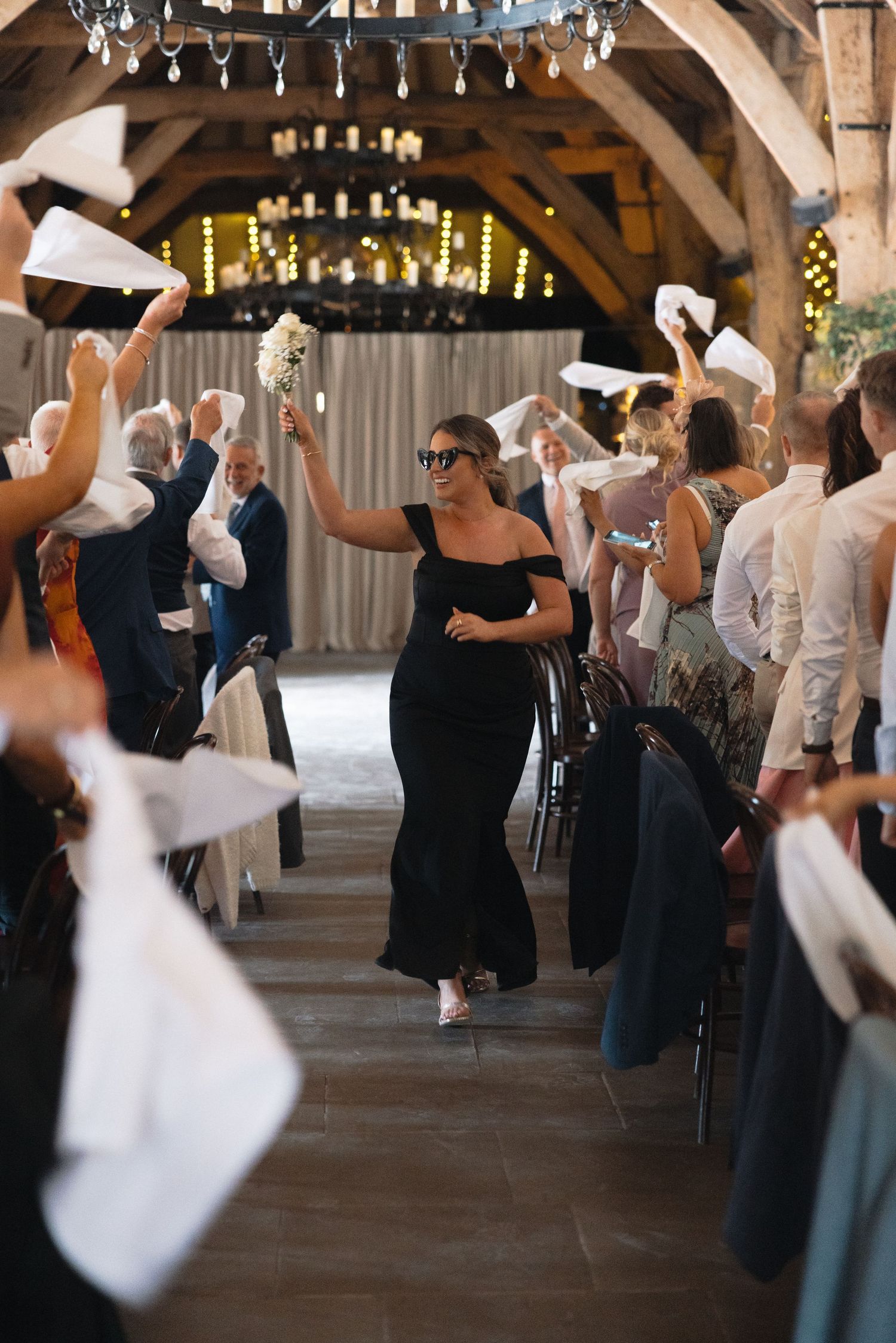 Wedding guests wave white napkins as someone walks down the aisle in a rustic venue.