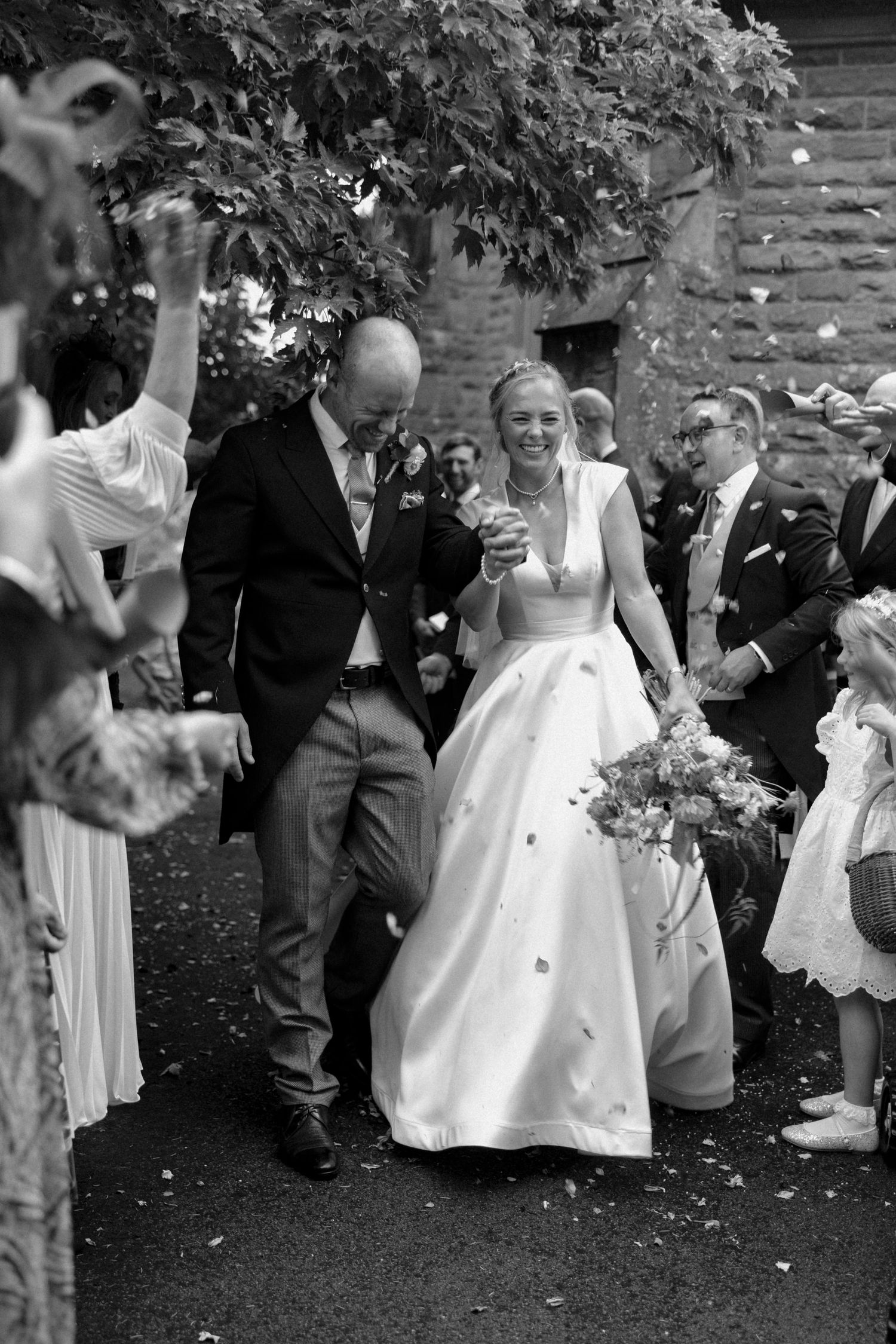 Wedding guests toss flower petals and confetti at a joyous outdoor celebration near a stone church.