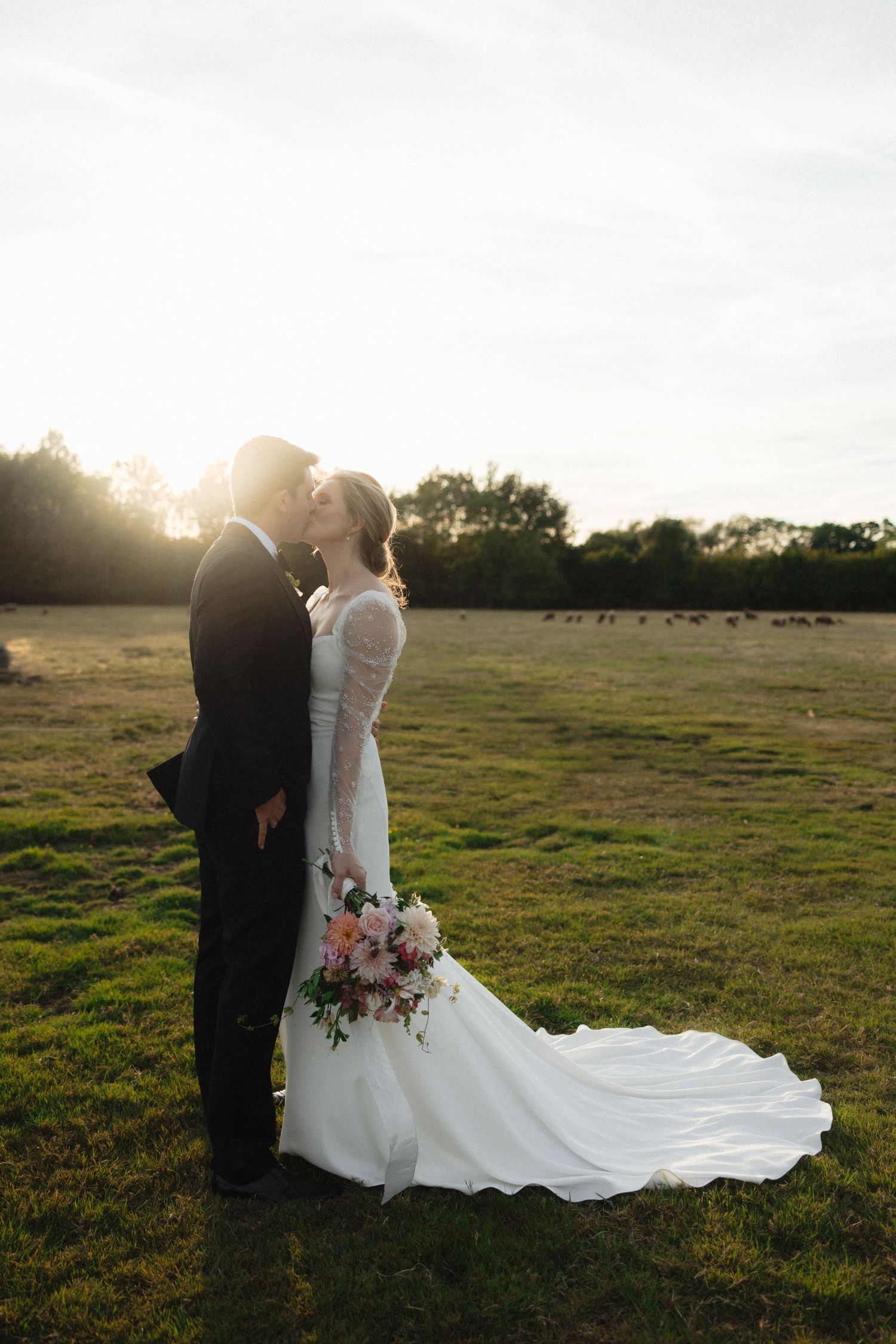 Wedding couple poses in field at sunset with flowing white dress train.