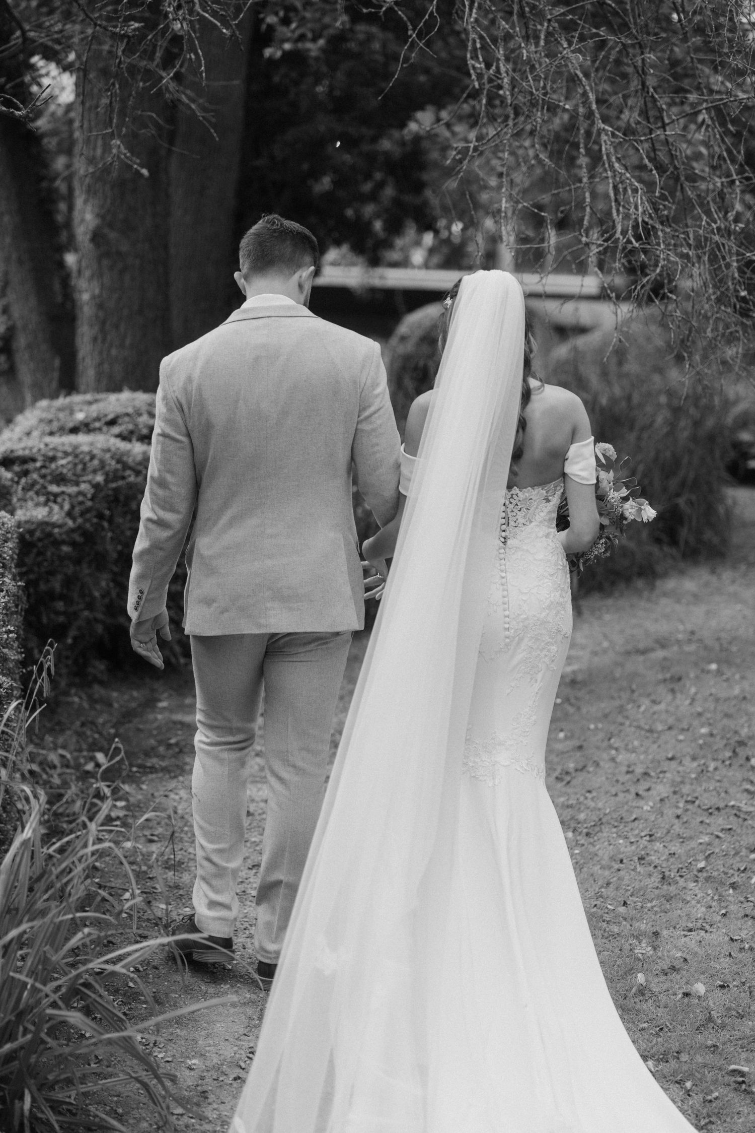 A couple in wedding attire walk along a garden path, with the bride's long veil trailing behind.
