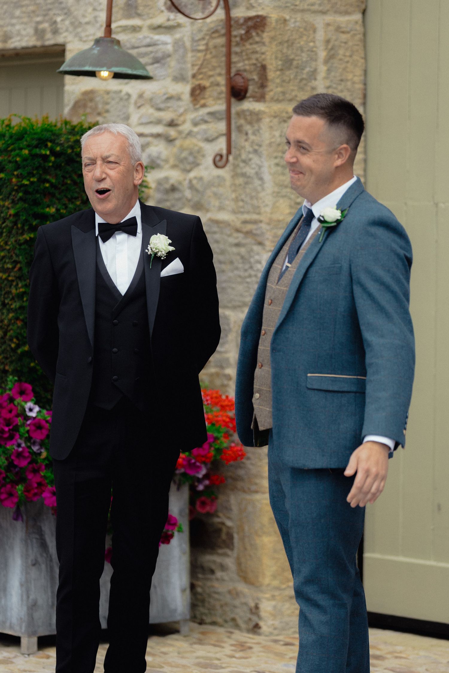 Men in formal attire share a lighthearted moment outside a stone building with flowering plants.