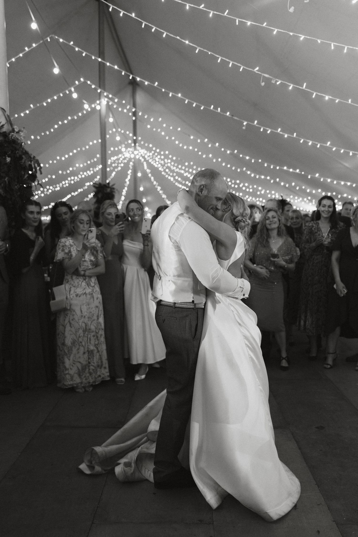 Black and white photo of wedding dance under string lights with guests watching in background.
