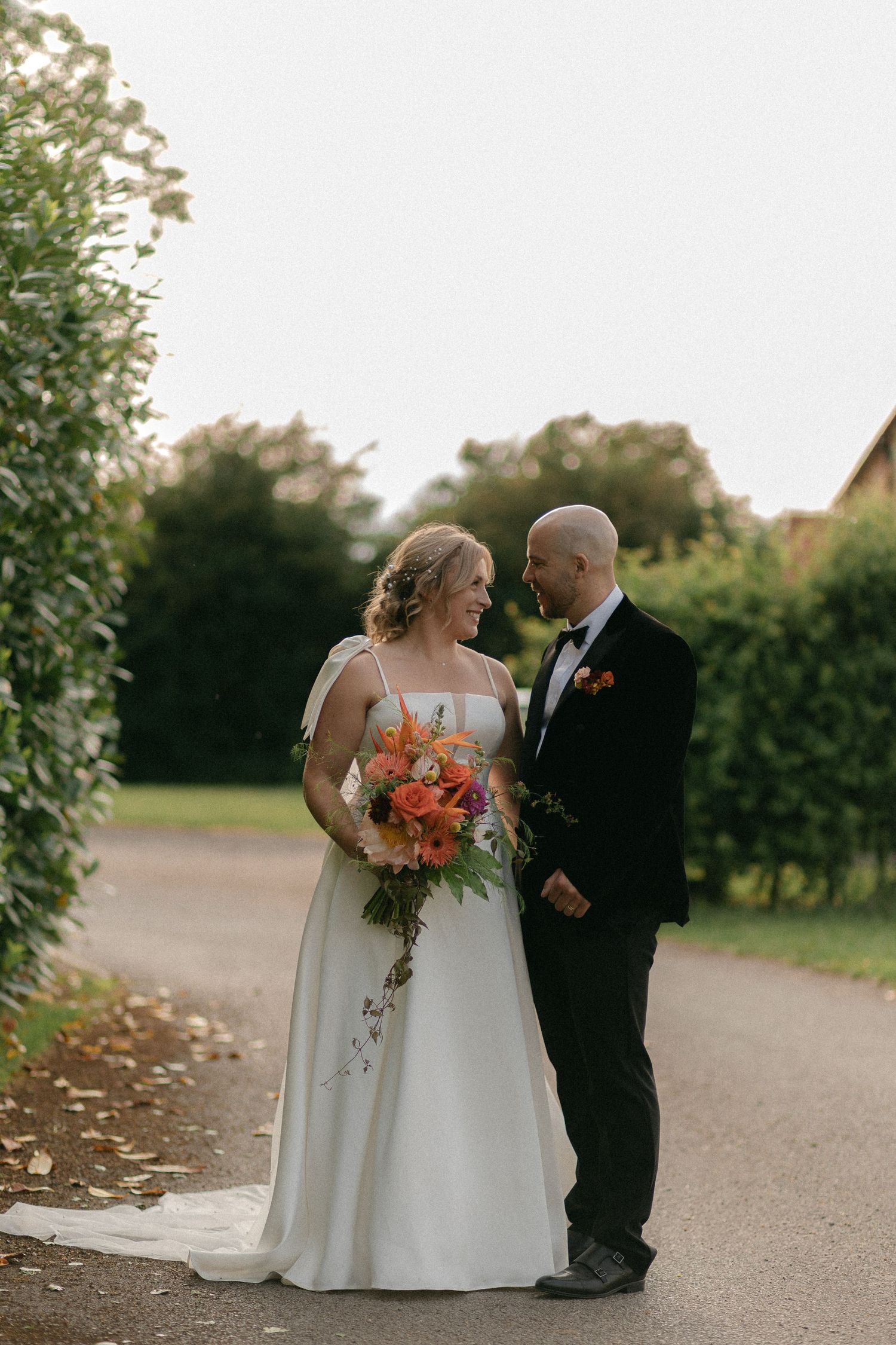 A wedding couple poses together on a path surrounded by greenery at sunset.