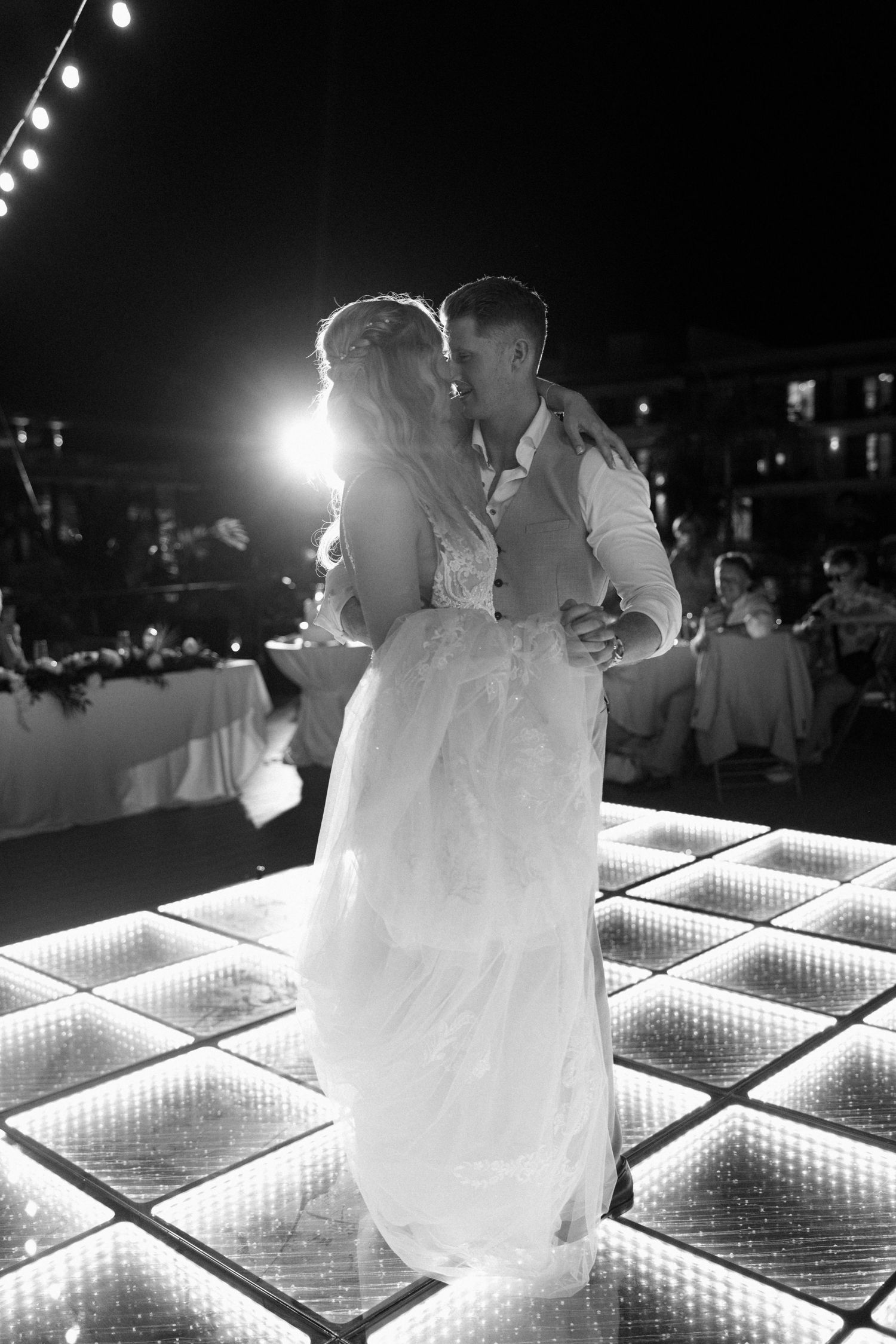 Couple shares an intimate dance on an illuminated dance floor under string lights at night.