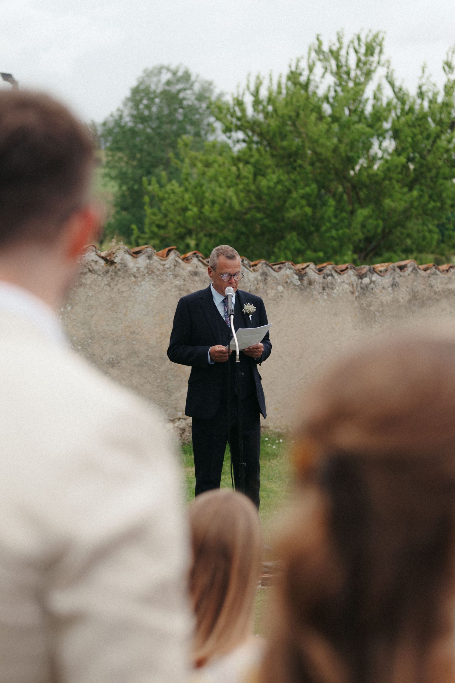 A speaker in a dark suit reads from papers during an outdoor ceremony with guests and greenery in the background.