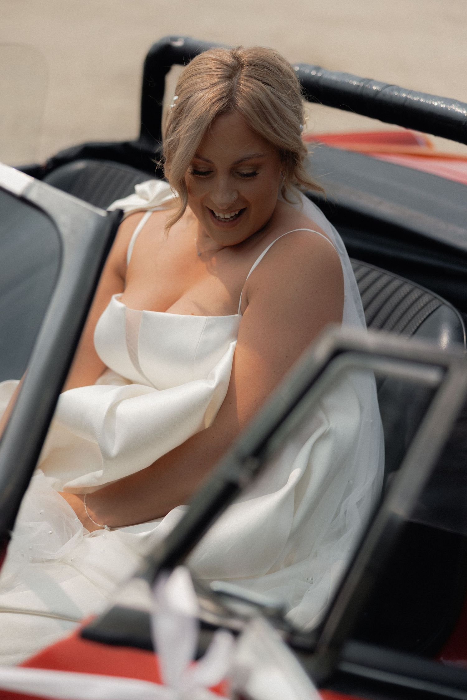 Woman in white wedding dress sitting in classic car with joyful expression.