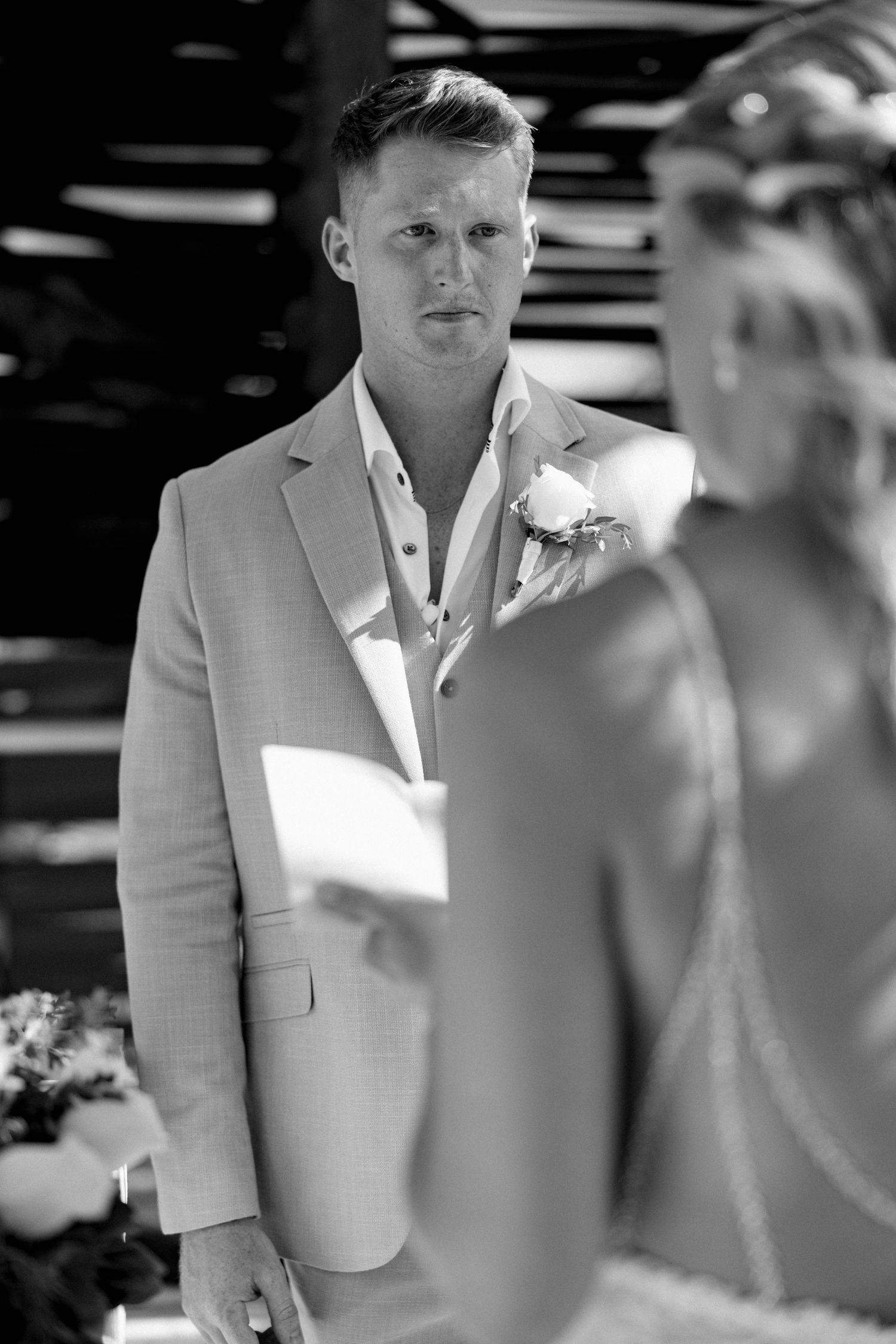 Groom in light colored suit stands attentively during outdoor wedding ceremony.