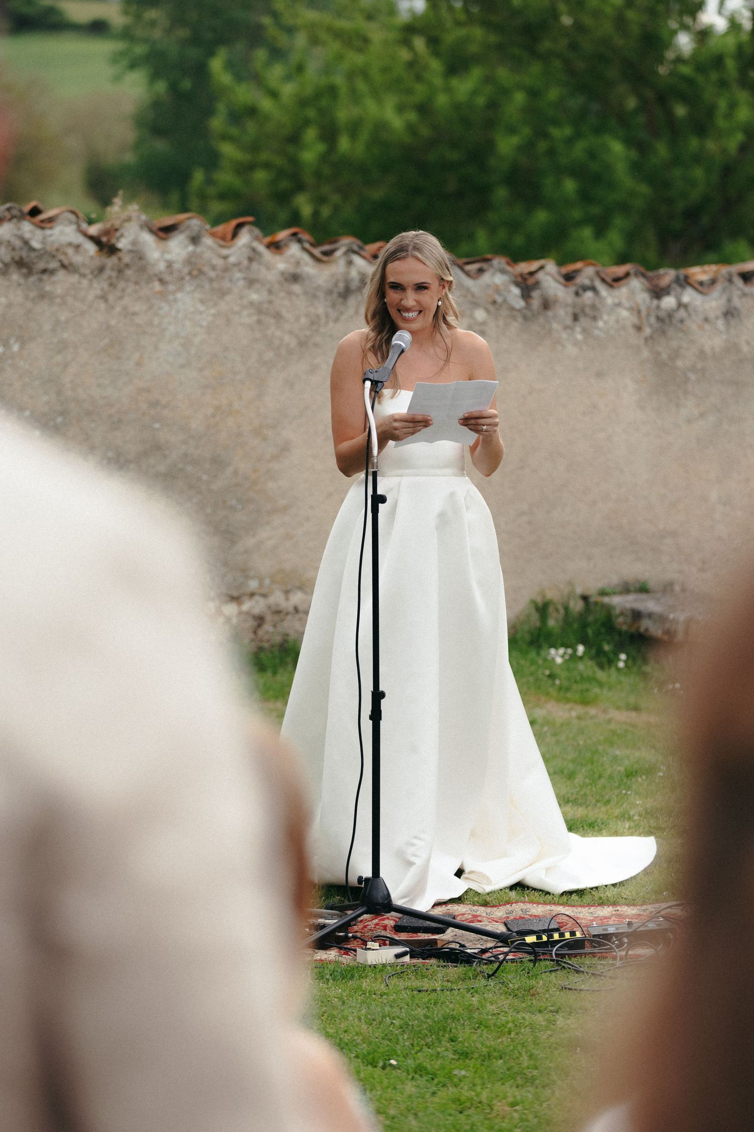 Bride in white gown standing near microphone setup against stone wall background.