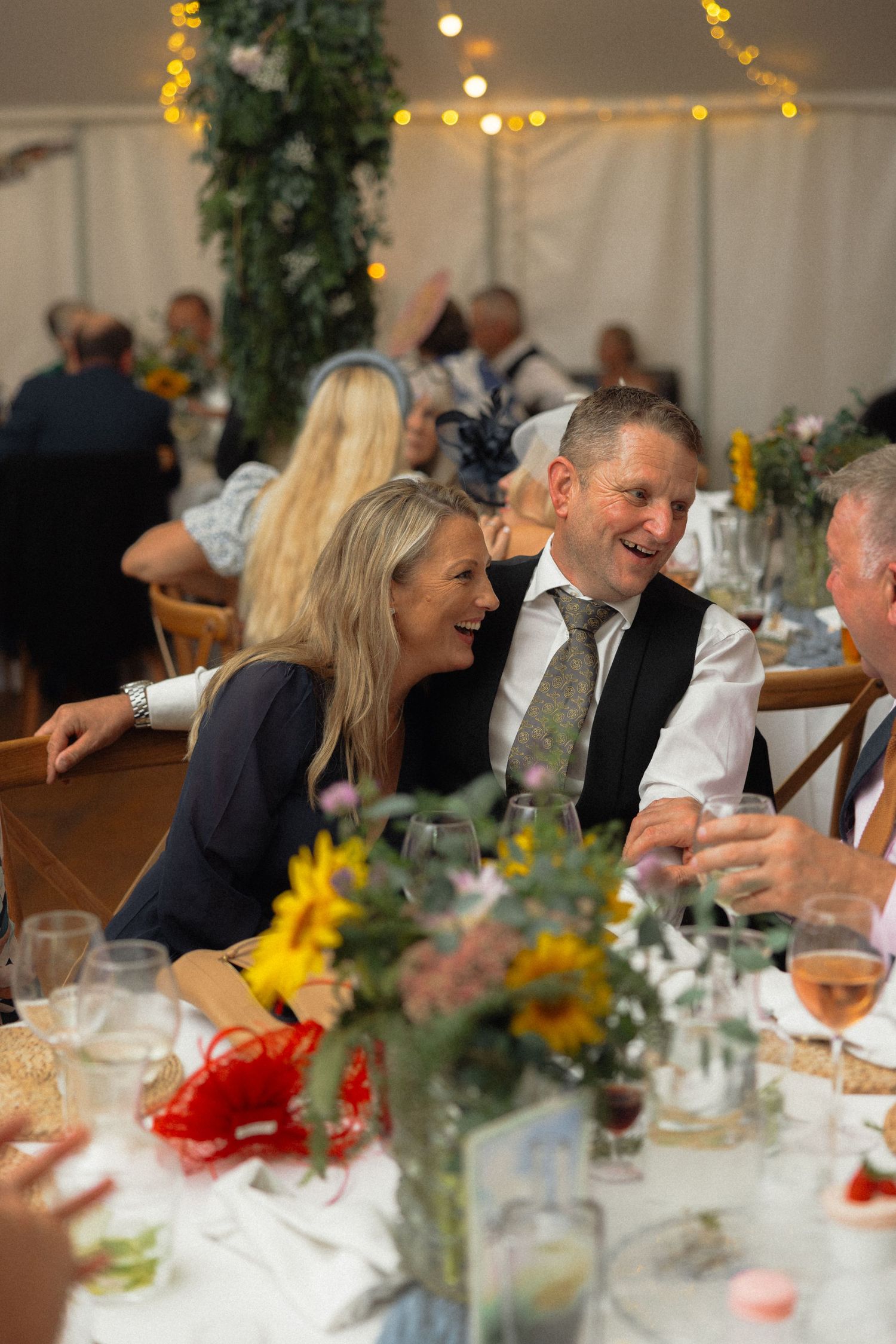 Wedding celebration with guests laughing at decorated tables adorned with yellow flowers and ambient lighting.