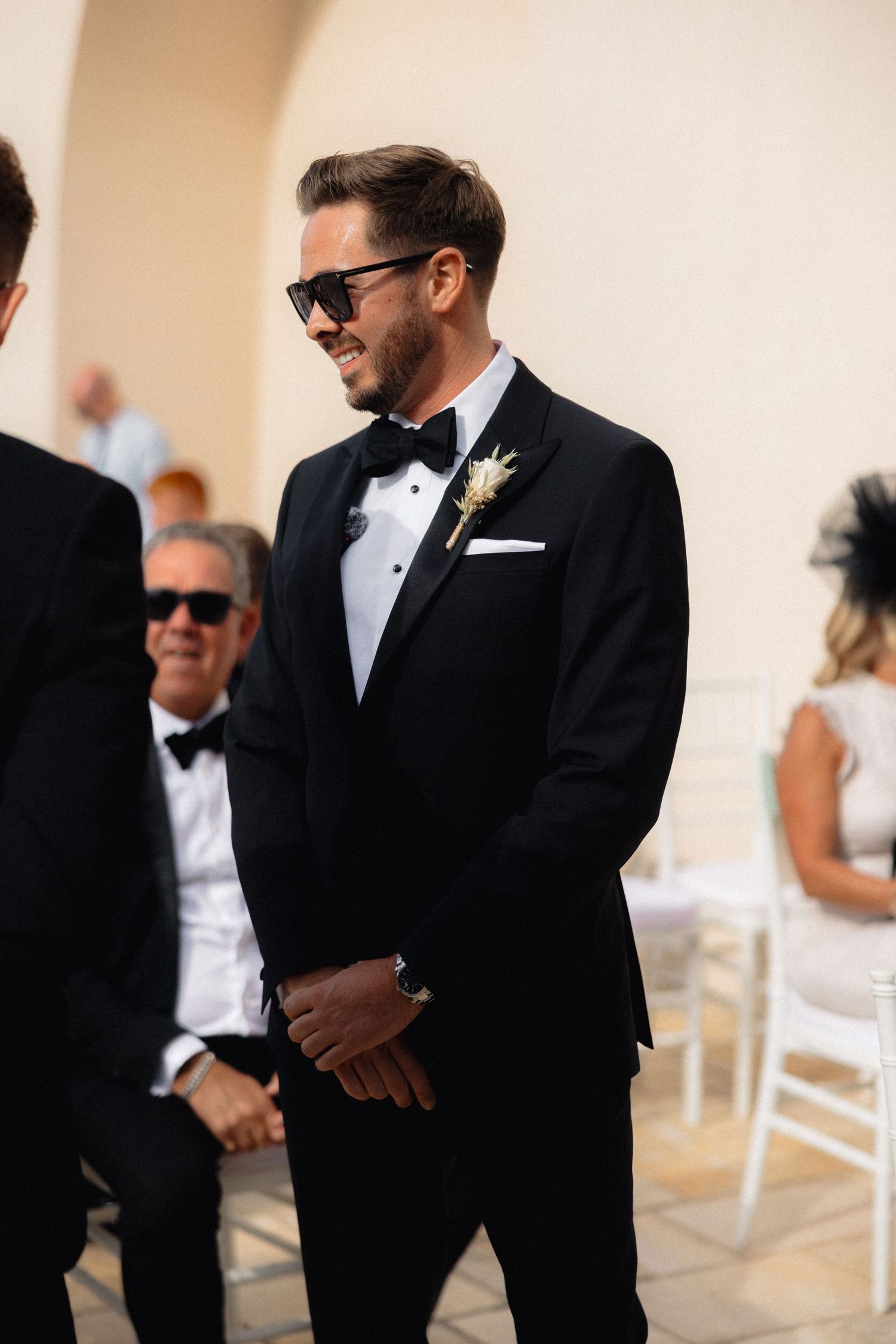 Wedding party members in formal black tuxedos gather during an outdoor ceremony.